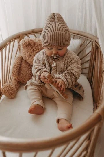 Baby in a brown knitted outfit and hat sitting in a wicker bassinet