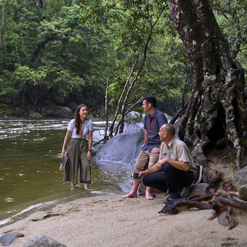 Tre persone conversano vicino a un fiume in una foresta. Una donna è in piedi nell'acqua bassa, mentre due uomini sono seduti su una radice di albero lì vicino.