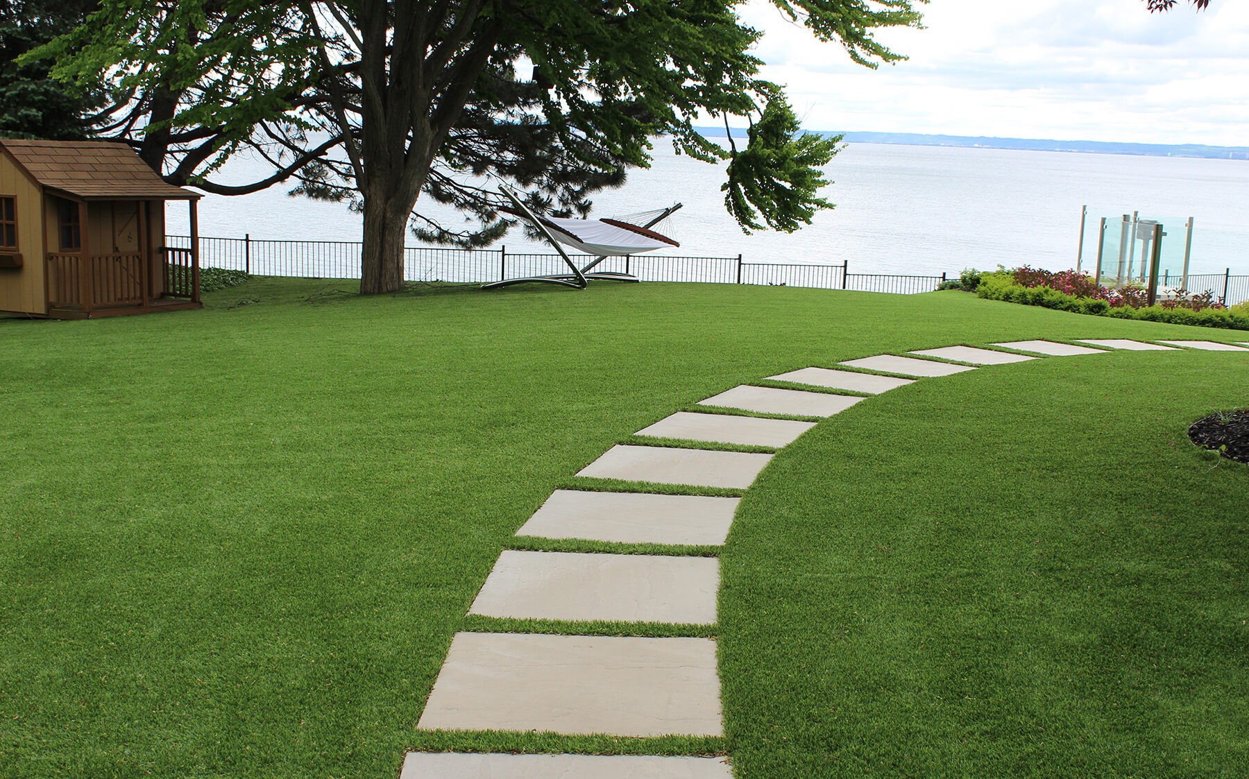 A lush green artificial lawn with a stone pathway that leads to a body of water and a small house and hammock nestled among the AGL Grass turf.