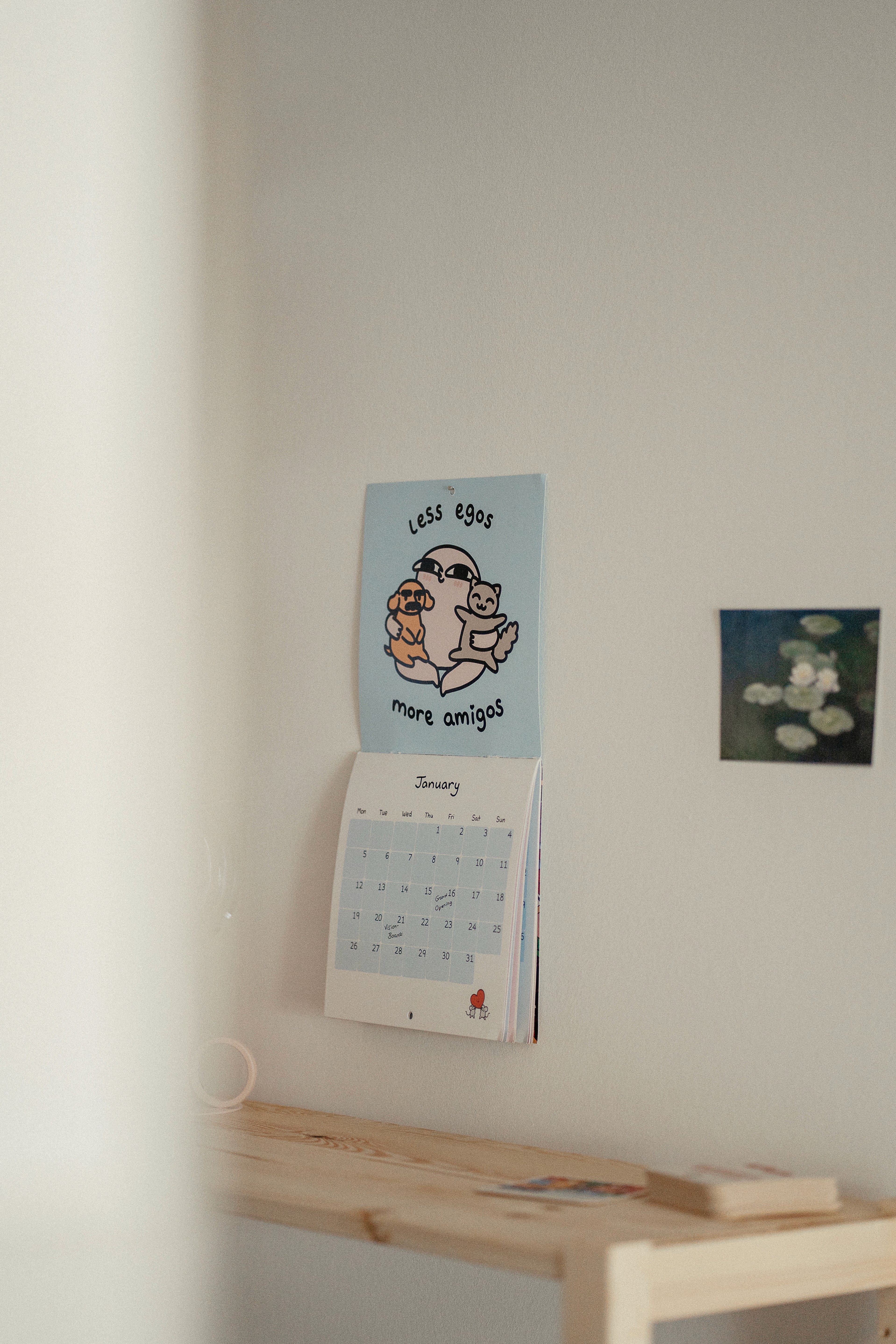 Wandkalender mit der Aufschrift 'less egos more amigos' und einer Zeichnung von drei Figuren über einem Holzregal in einem Coworking Space.