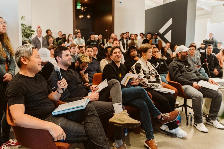 A diverse group of people sitting and standing in a large, well-lit room, attentively listening to a speaker. The foreground shows a few individuals sitting in chairs, smiling and taking notes, with one person holding a microphone. The background is filled with a crowd, some seated and some standing, indicating an engaged and lively audience. The atmosphere is collaborative and focused, suggesting a workshop or presentation setting.