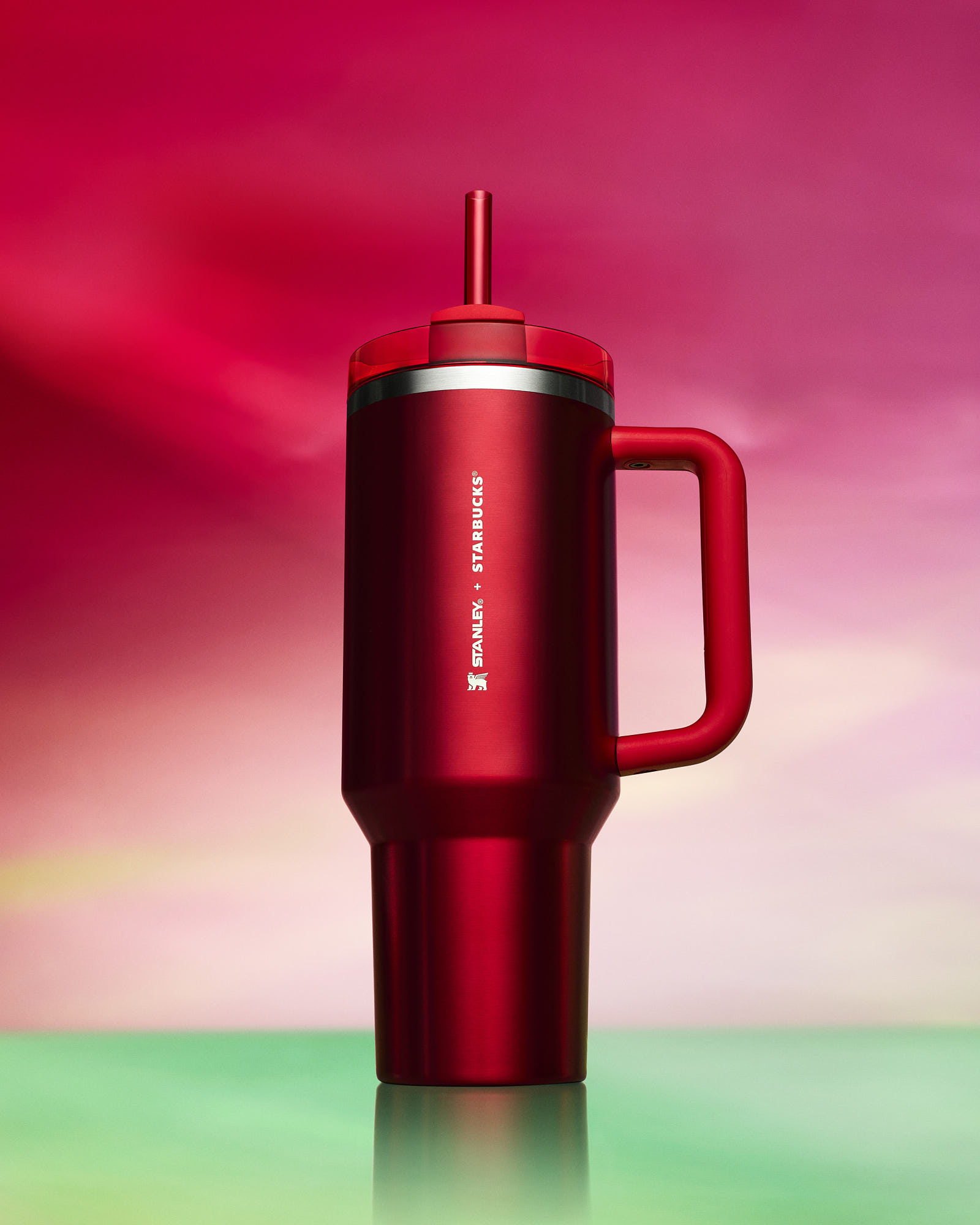 A tall red stanley tumbler with a handle, set against a gradient pink and green background.