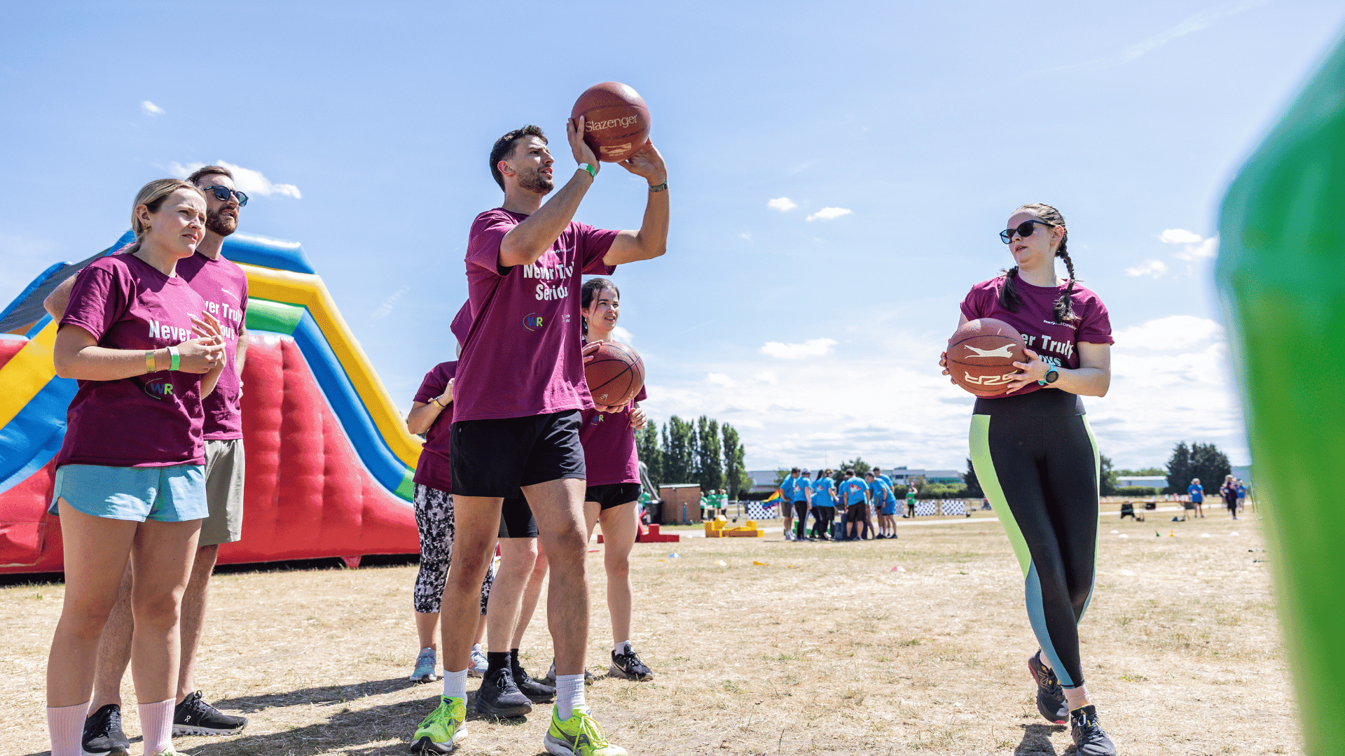 Outdoor team-building event with participants wearing matching blue shirts, taking part in inflatable obstacle courses and games, smiling and cheering, with the words 'ENTER A TEAM' in large bold white and green text across the image.