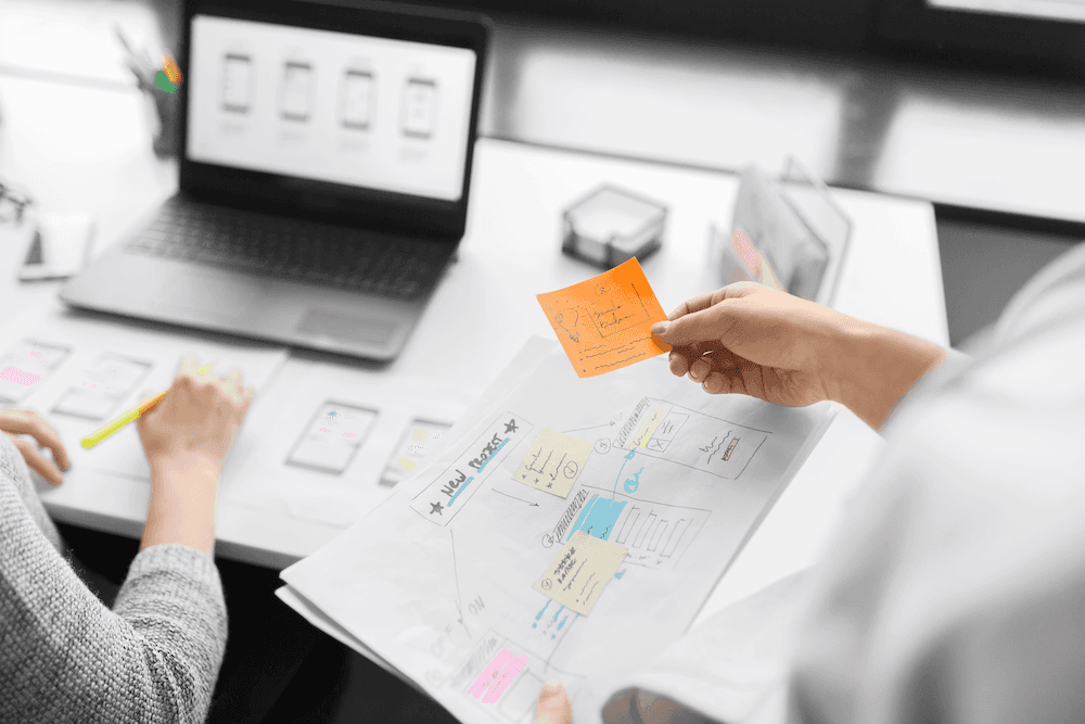 Hand hält orangefarbenen Notizzettel über einem Papier mit Skizzen für Benutzeroberflächen, im Hintergrund Laptop und weitere Papiere auf einem Tisch