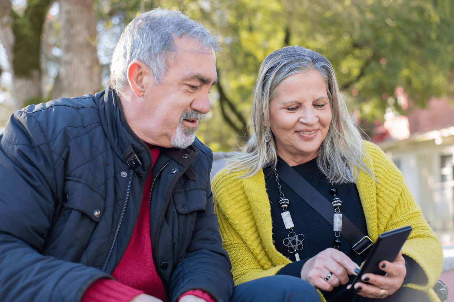 An older man and woman sitting outdoors in the sun, smiling at a phone screen they are looking at together.