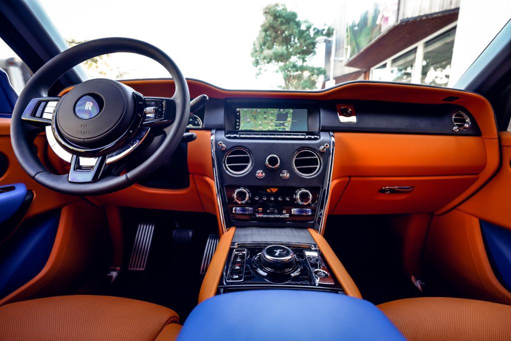 Interior dashboard view of Rolls-Royce Cullinan showing orange leather upholstery, digital displays, and premium wood trim details