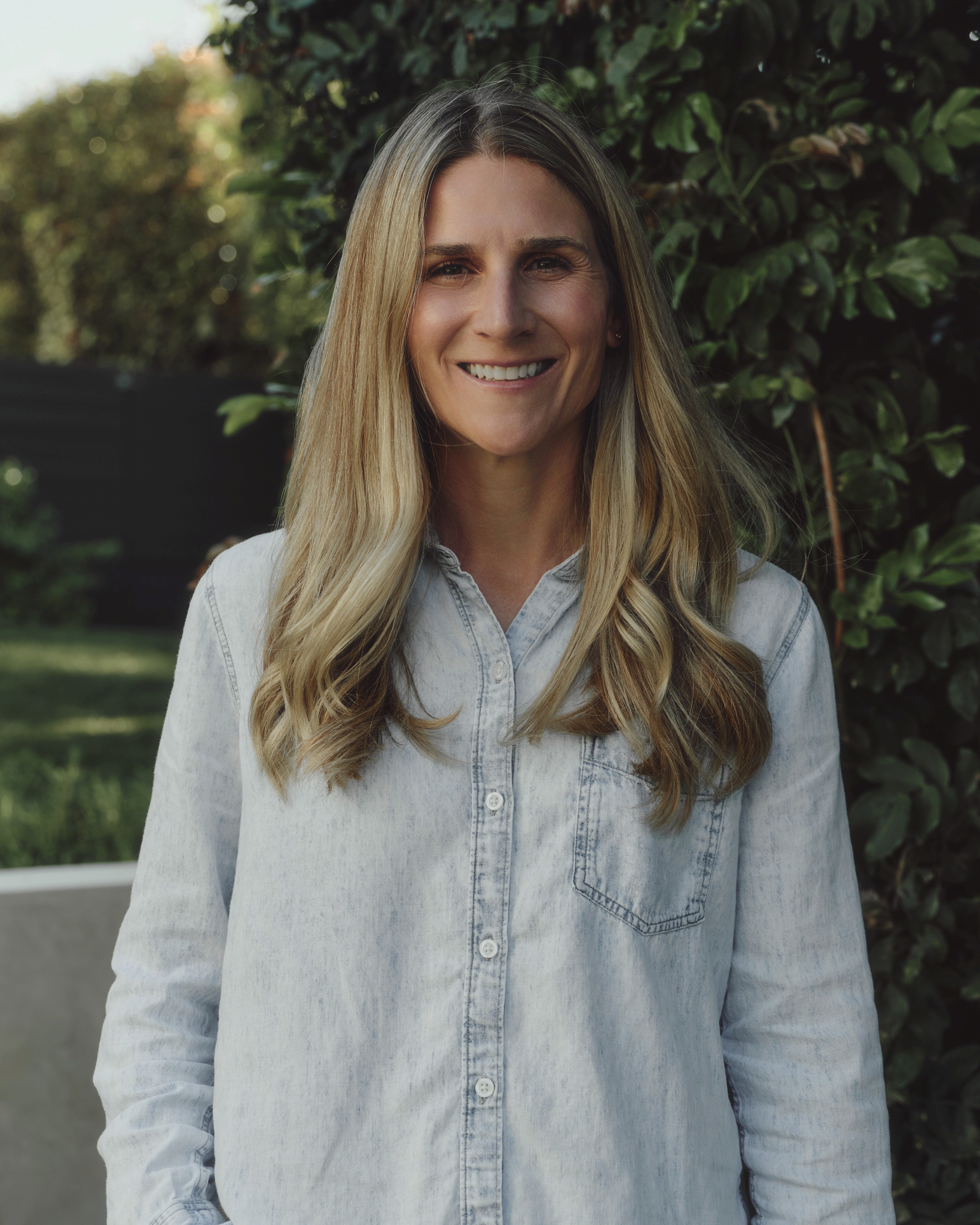 A professional portrait of a smiling woman with long blonde hair, wearing a denim button-down shirt, standing in front of lush green foliage