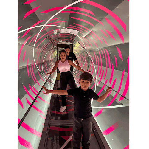三個人走過一個旋轉的隧道，隧道裡有粉紅色和白色的條紋，緊握著扶手以保持平衡。