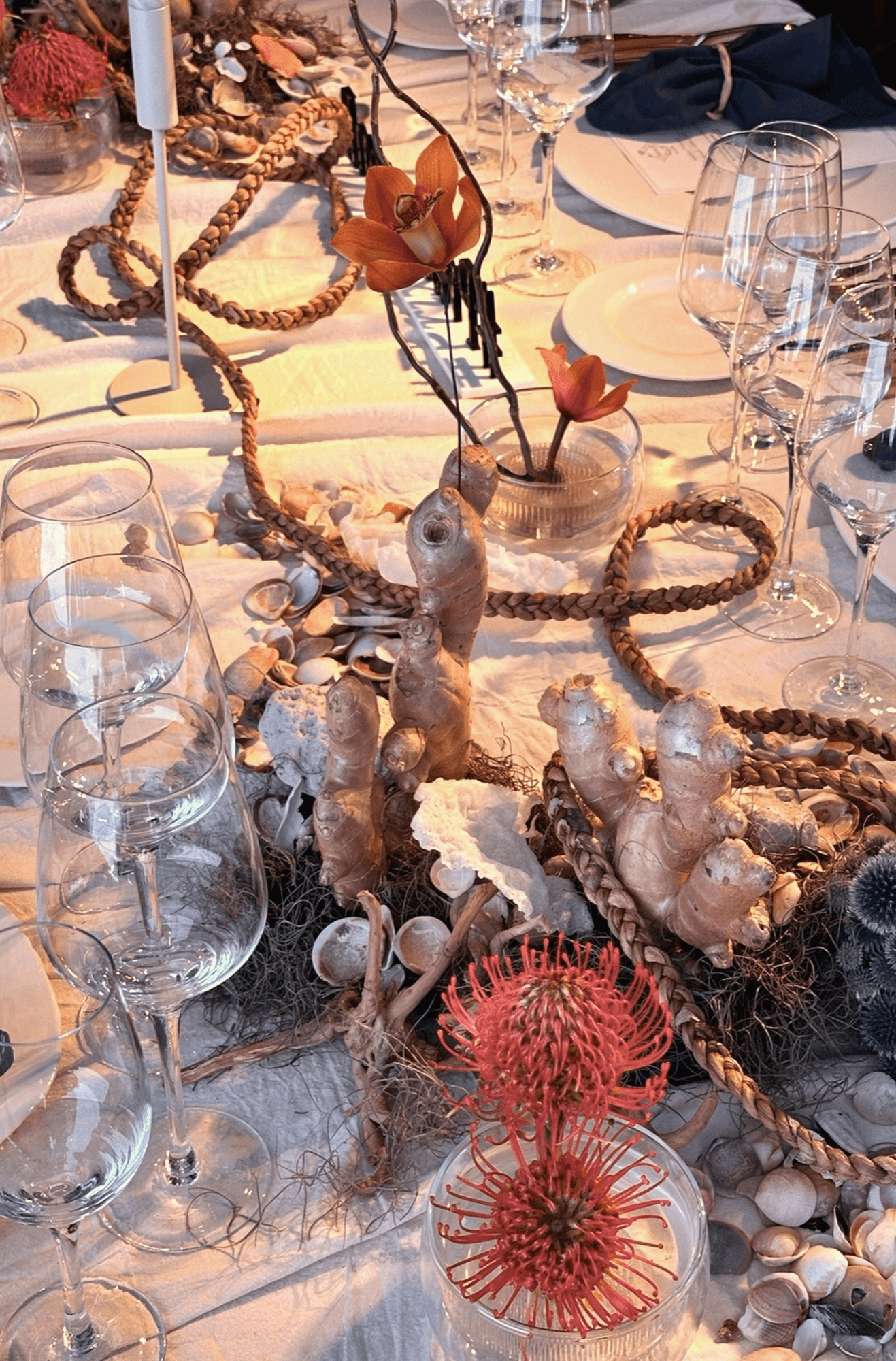 Centro de mesa floral con hortensias rosadas, flores blancas y amaranto rojo, acompañado de una composición de vegetales frescos como maíz, pimientos y chauchas, sobre un mantel blanco ilustrado, en un salón de estilo clásico con paredes decoradas