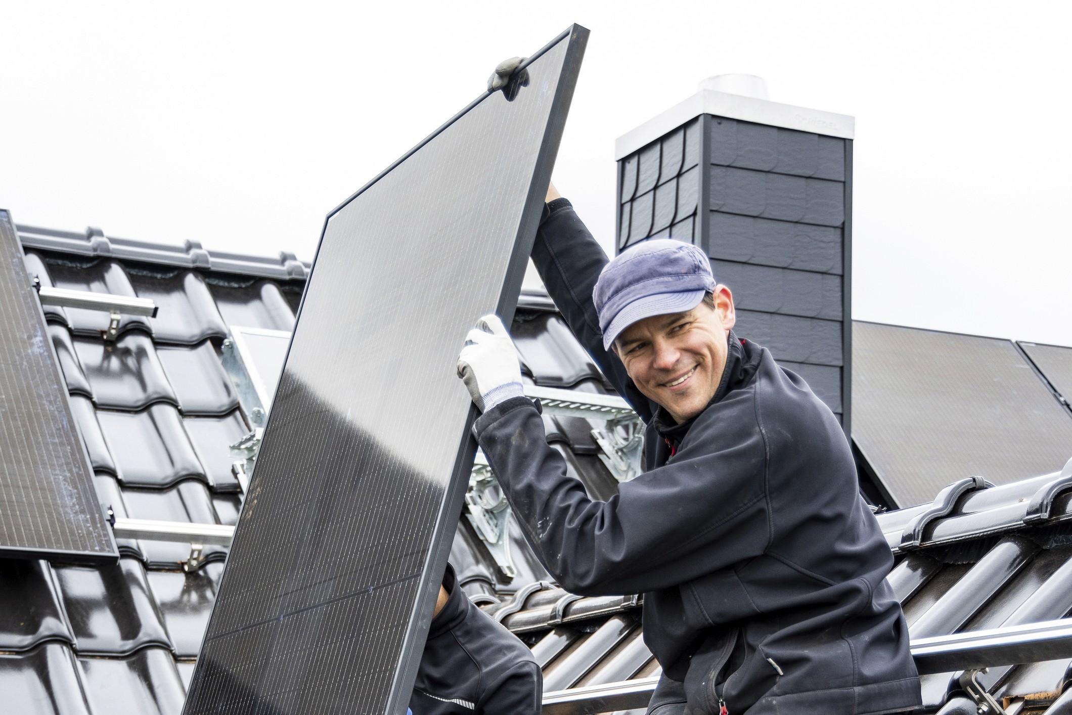 Ein Arbeiter mit Hut posiert mit einem großen Solarpanel, im Hintergrund ist ein modernes Dach zu sehen.