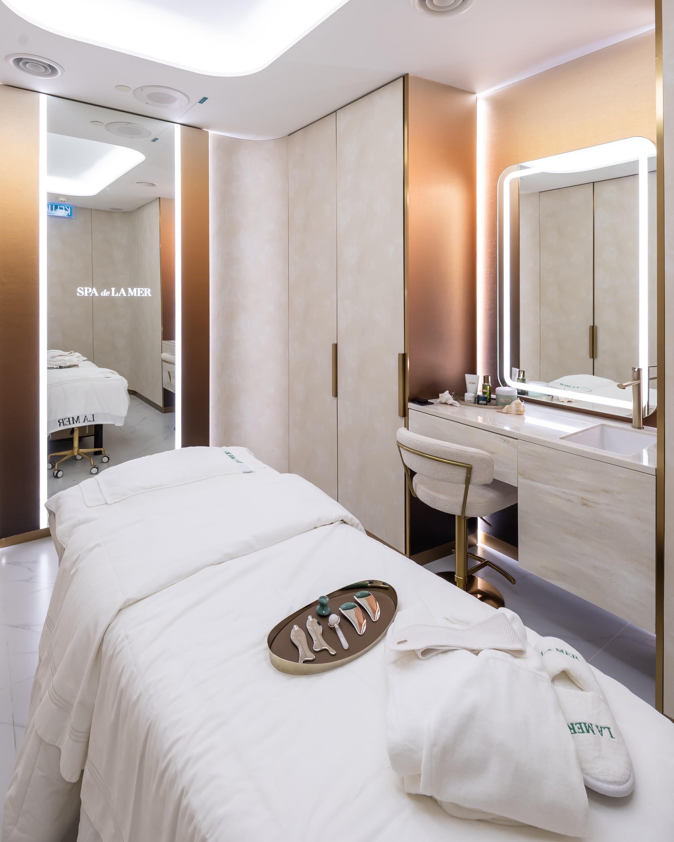 A luxurious spa treatment room featuring a massage table with white linens, a tray of skincare tools and products, and a sleek vanity with a mirror. The room has warm lighting and elegant decor.