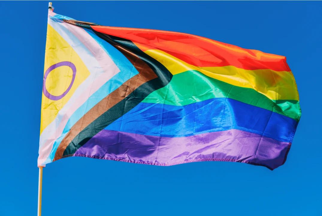 Uma vibrante bandeira do Orgulho Progressista, com listras do arco-íris e cores adicionais que representam comunidades marginalizadas, incluindo preto, marrom, azul-claro, rosa e branco, tremula contra um céu azul limpo.