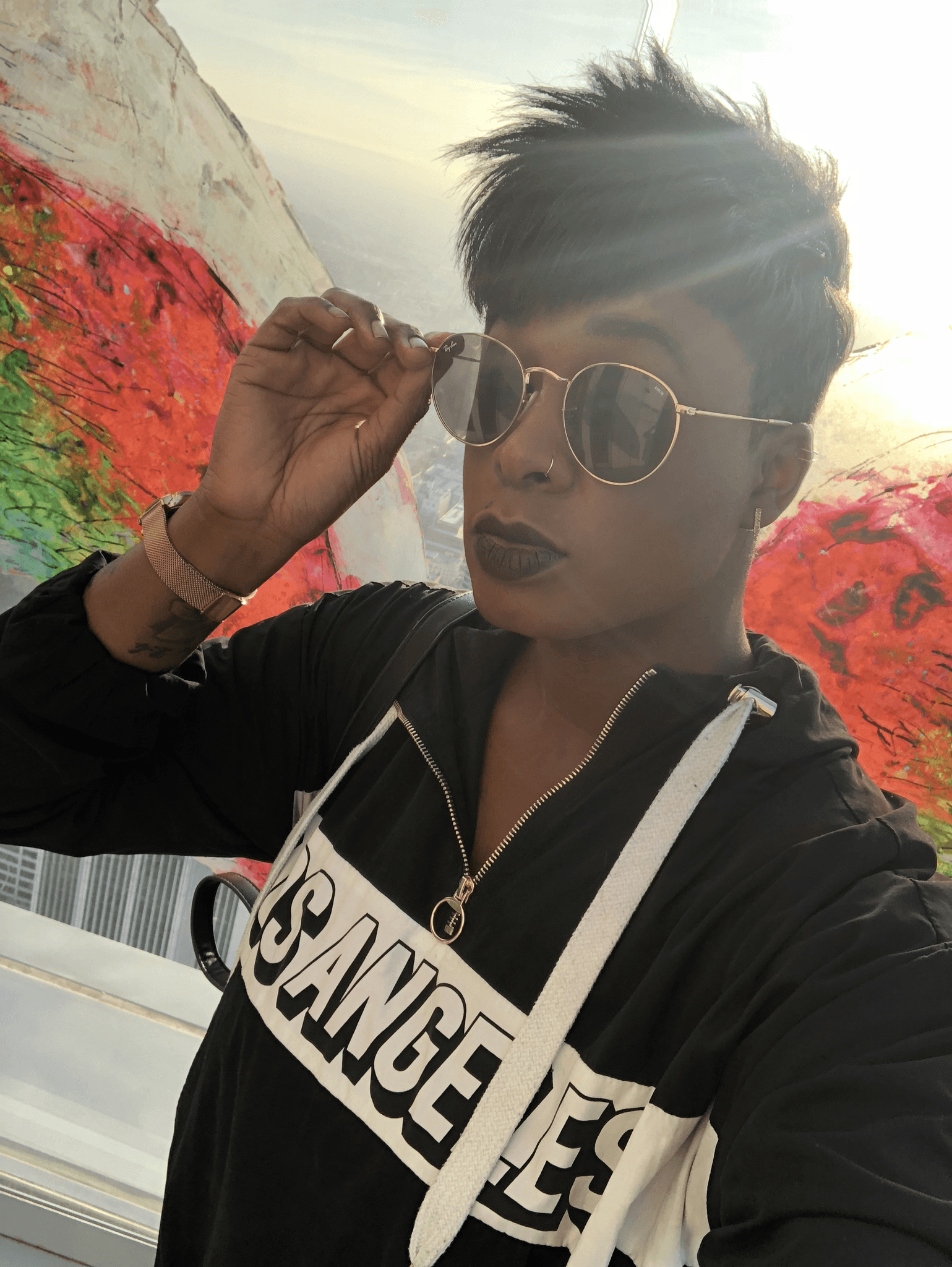 Ashleigh Warren wearing sunglasses and a black hoodie poses outdoors with colorful flowers in the background.