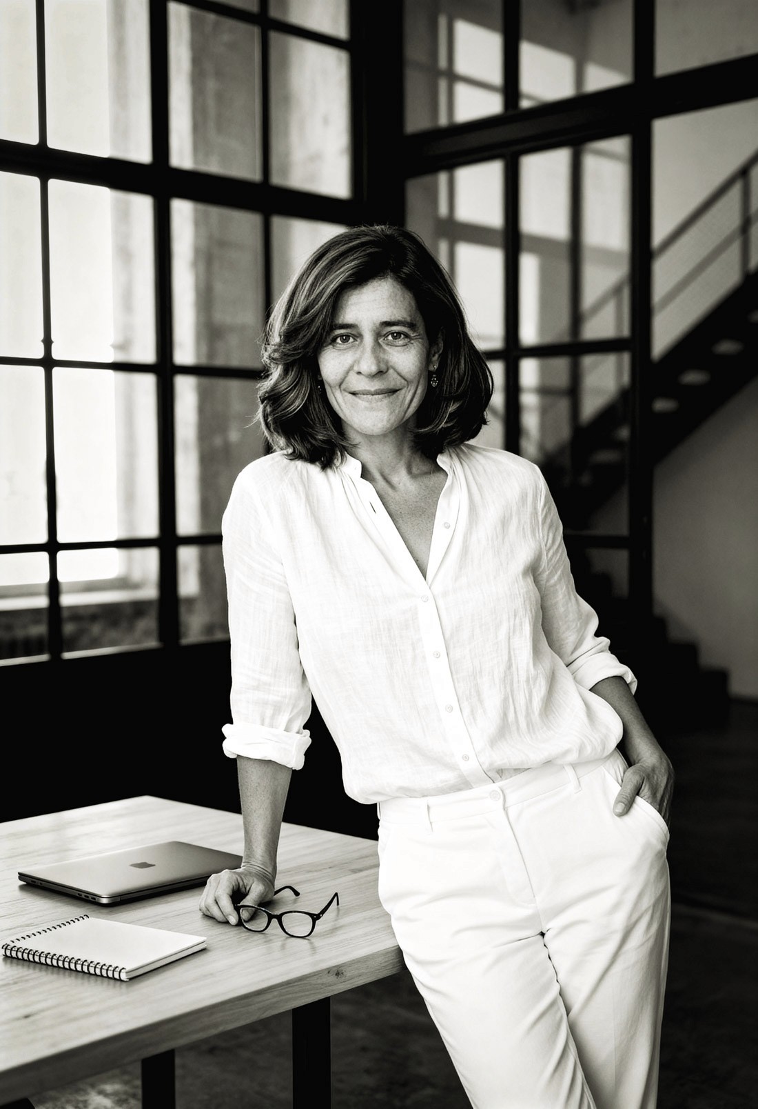A woman in a white blouse stands confidently at a table, framed by large windows in a stylish interior.