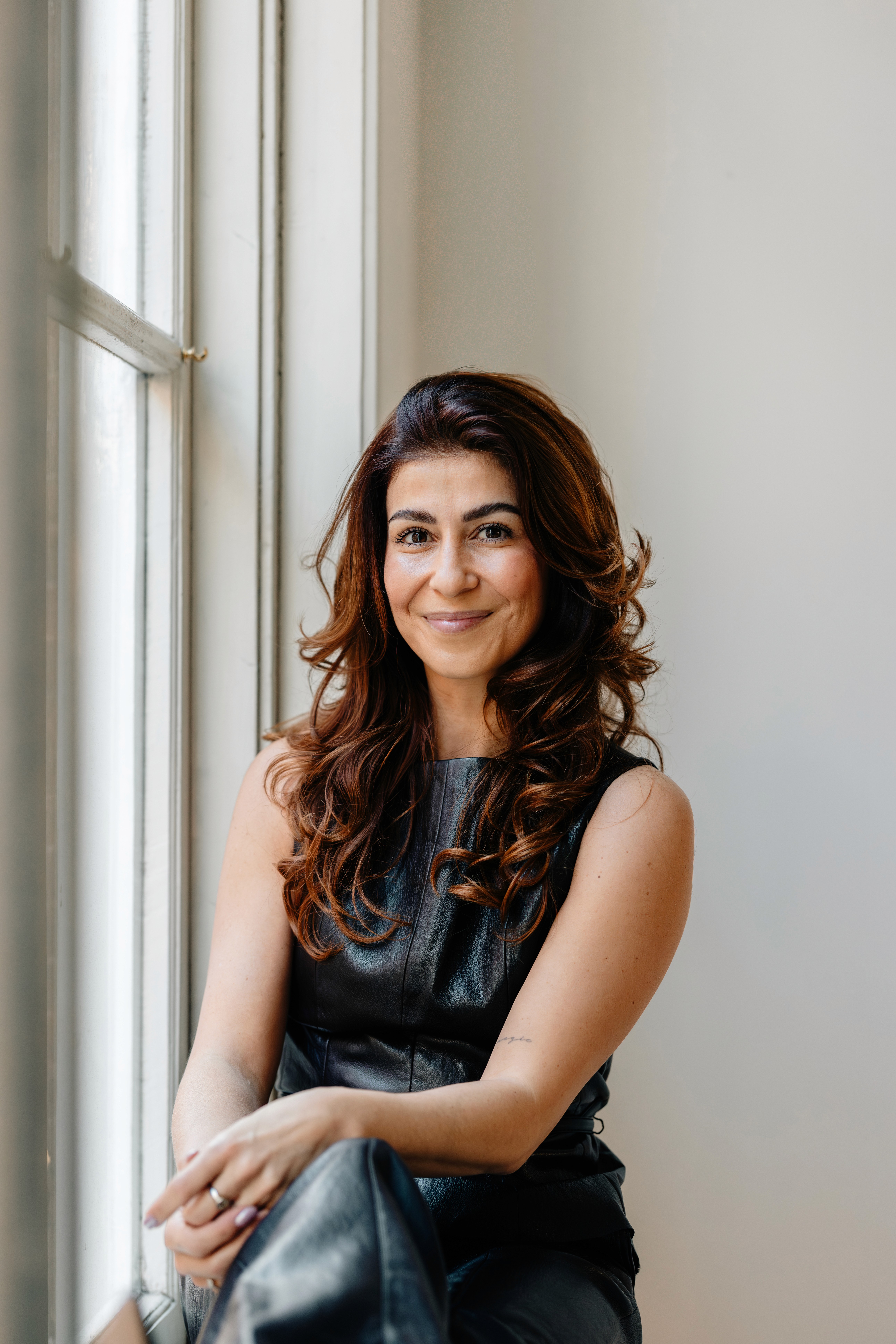 A woman with long, curly hair sits by a large window, smiling and wearing a black top and blue jeans.