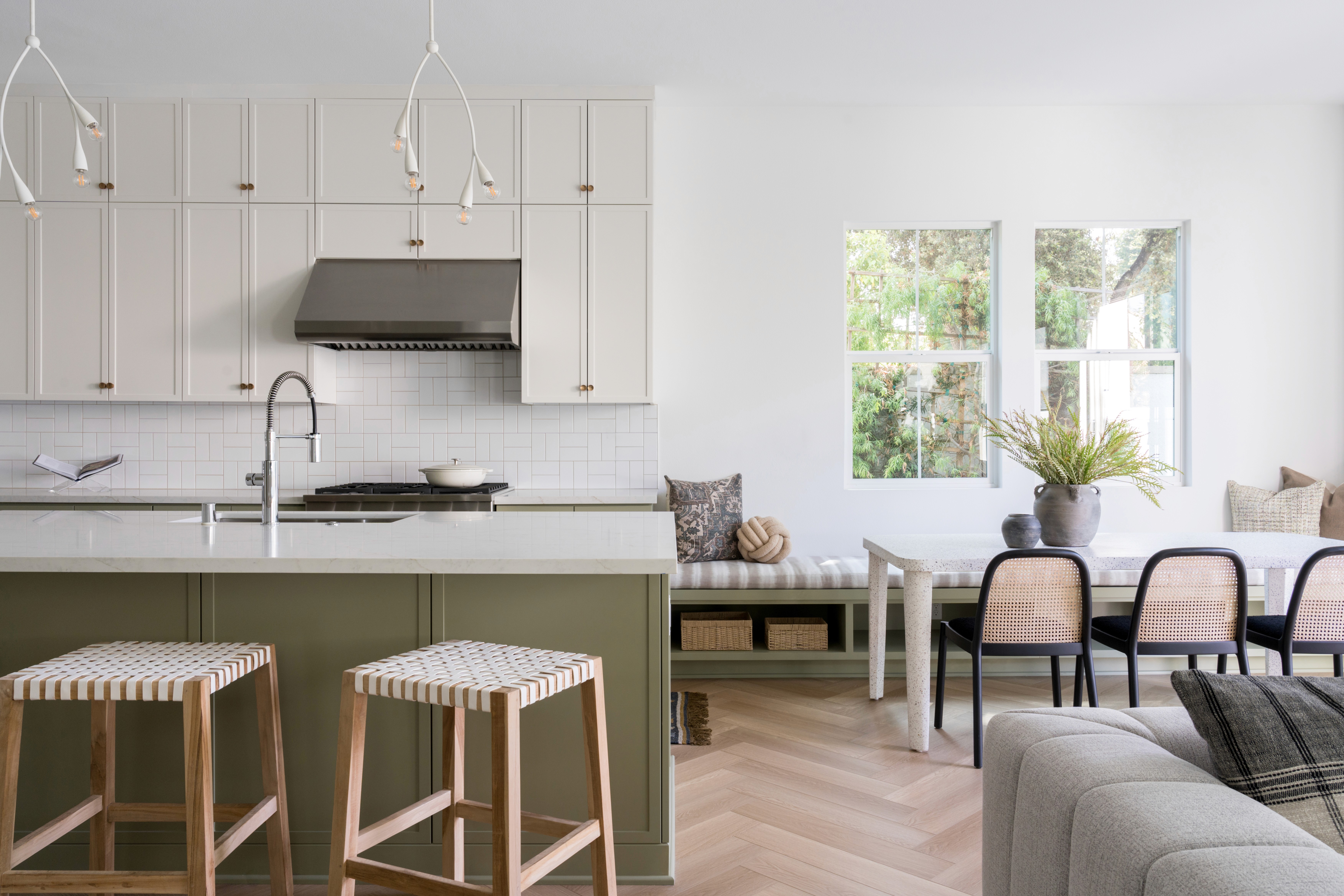 Fresh, open kitchen and seating area with sage green paneled island, white countertops, chrome faucet, chevron wood flooring, white cabinets with brass finishes, light wood backless barstools, built-in bench seating with white and grey cushions, white dining table,  and black and rattan chairs by Jubilee Interiors in Arcadia, California