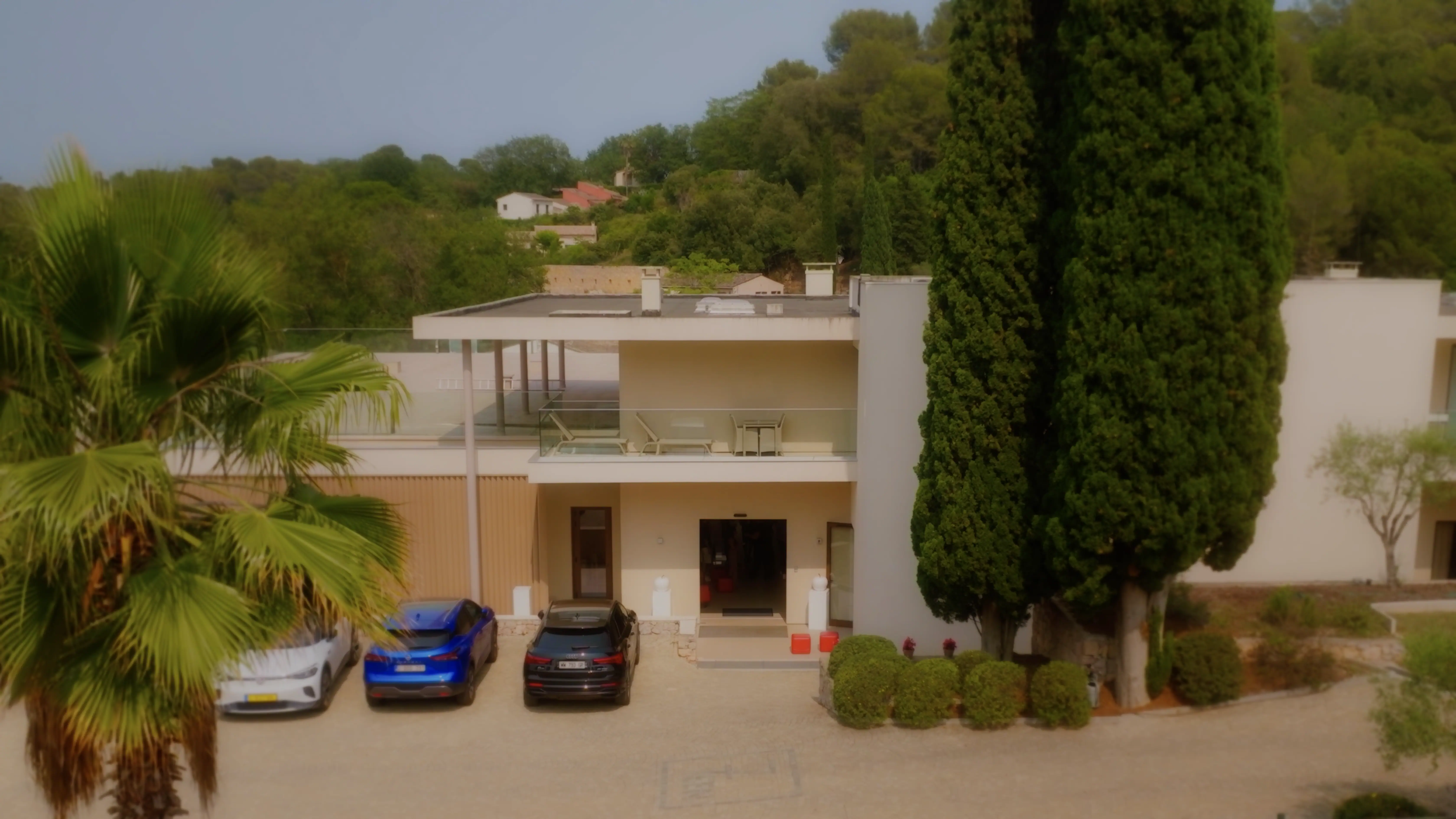 A modern villa surrounded by lush greenery features a sleek, two-story design with large windows and a spacious balcony, while three luxury cars are parked in the driveway, shaded by tall, elegant trees.