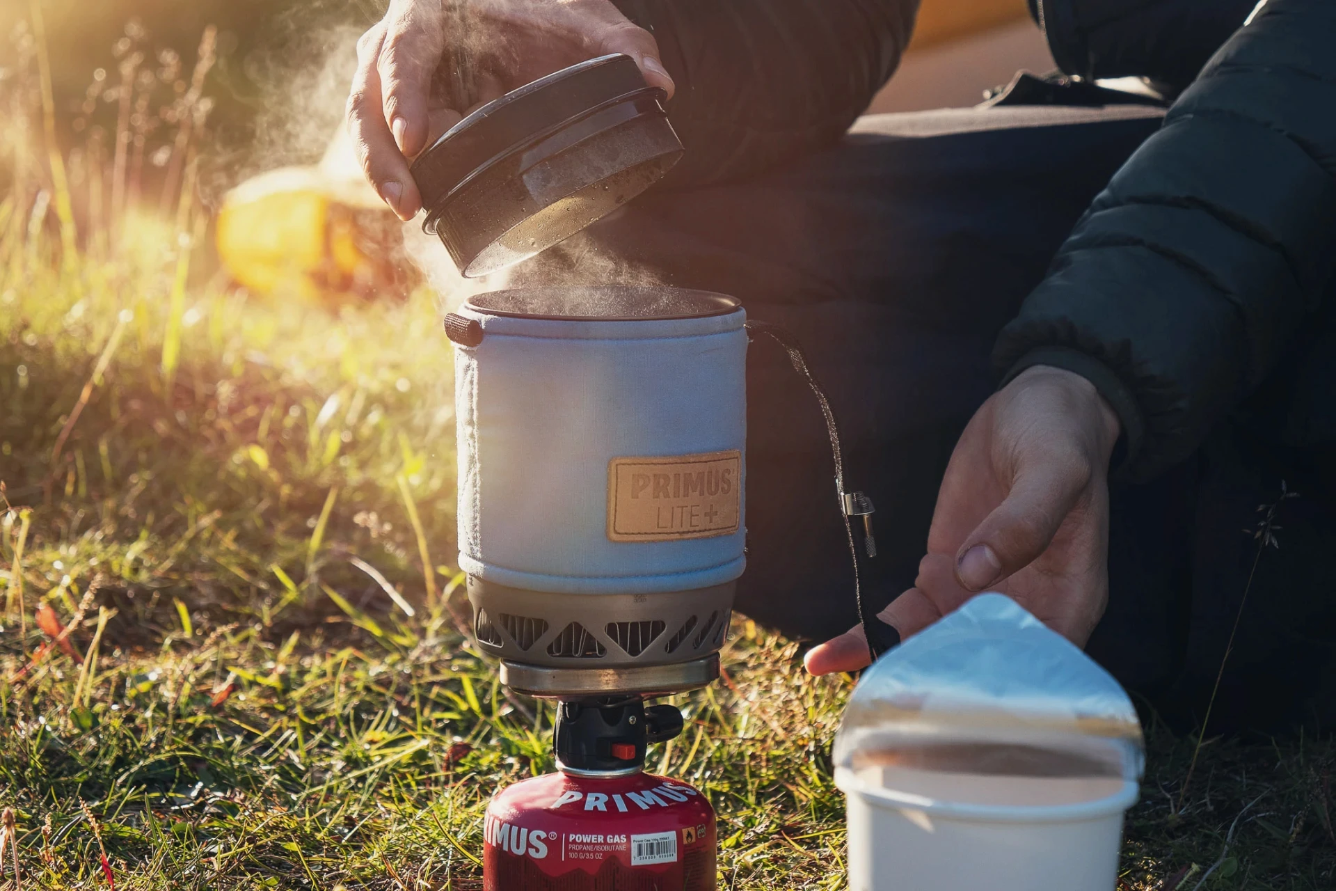 Hands cooking on a small gas stove outdoors, steam rising in sunlight.