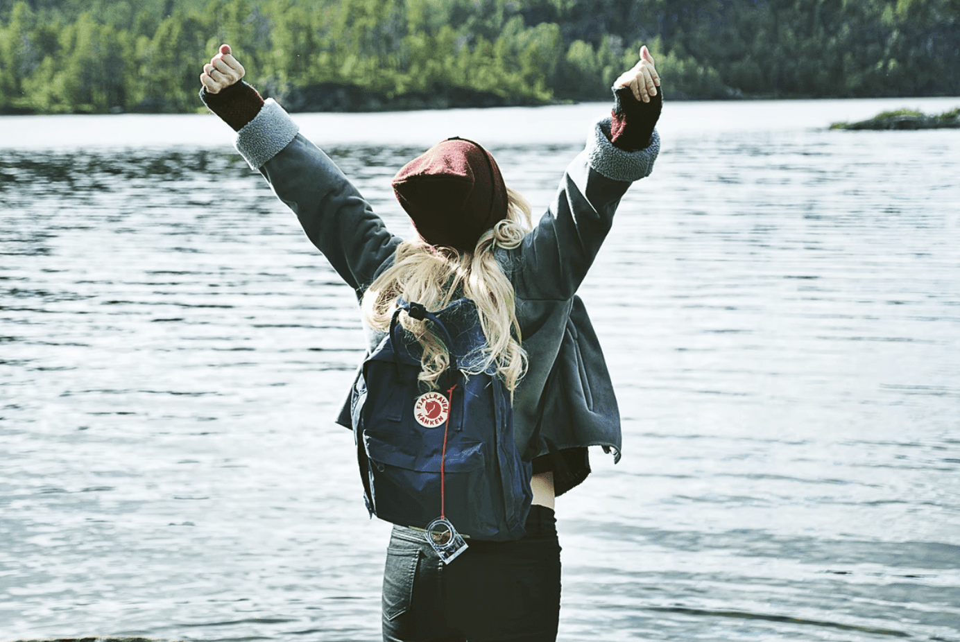 A person with long hair wearing a backpack stands by a lake, arms raised in excitement or celebration.