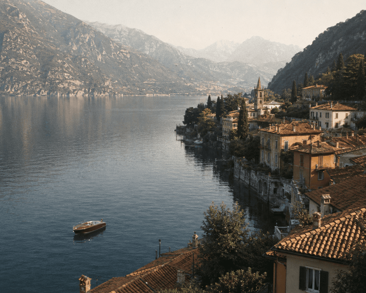 A scenic high-angle photograph of a historic coastal village along Lake Como in Italy. Terra-cotta roofed stone buildings with shuttered windows and a central church steeple cluster along the steep, lush shoreline. A classic mahogany wooden speedboat drifts on the calm, deep blue water, surrounded by dramatic mountain slopes fading into the soft haze of the distance.