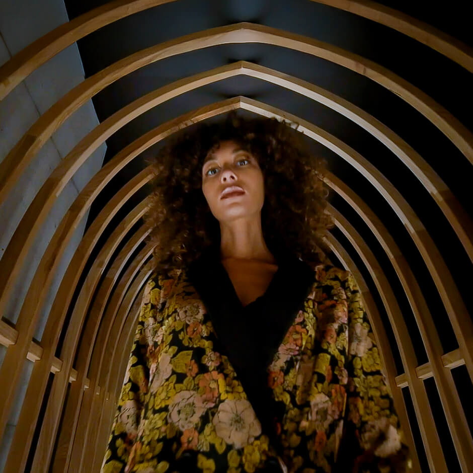 A woman with curly hair stands under a wooden arch structure, wearing a floral patterned jacket and looking straight ahead.