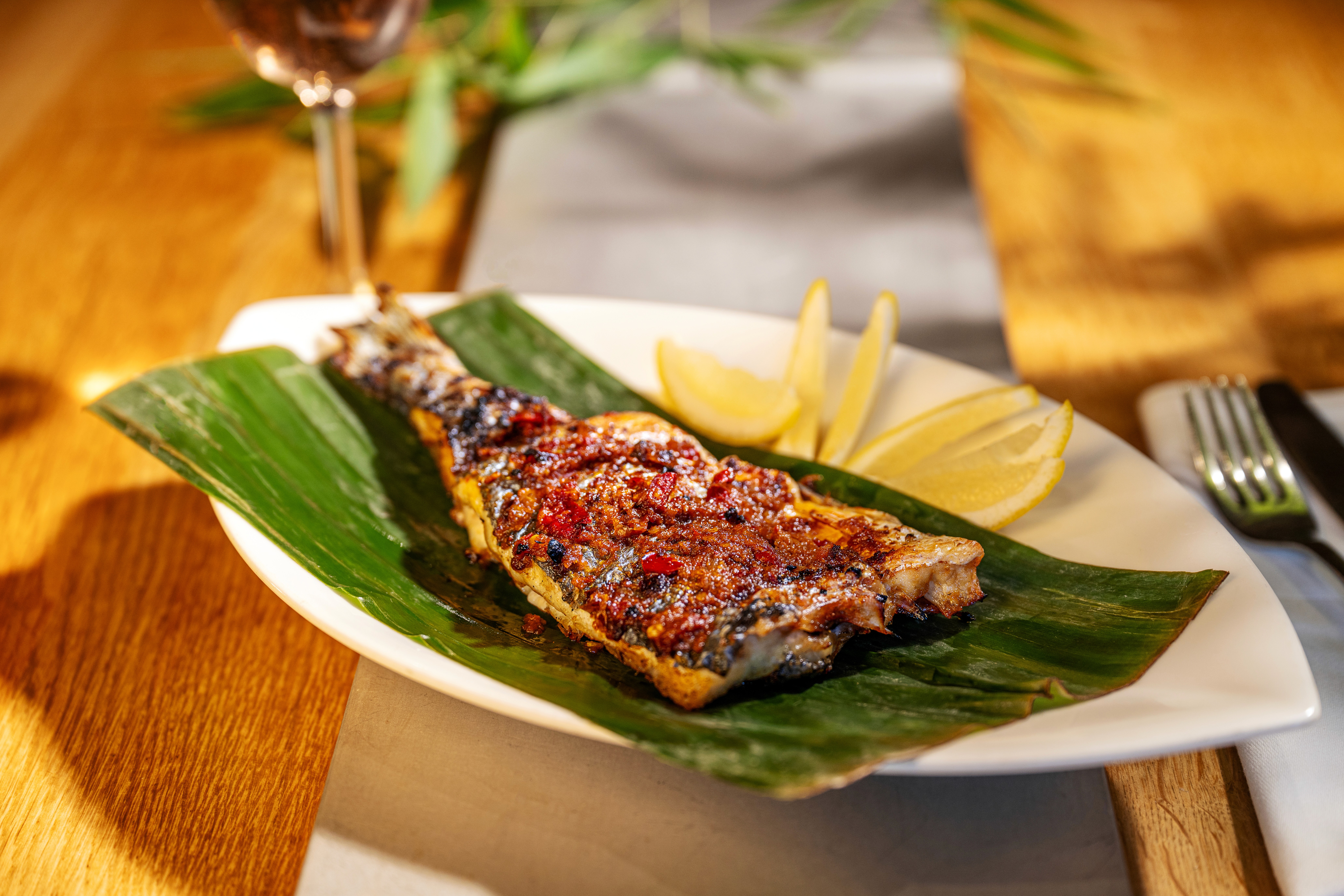 Close-up van Authentieke Ikan Bakar Jimbaran: een hele rode snapper, rijkelijk bestreken met rode sambalmarinade, perfect gegrild op kokosbriketten en geserveerd met partjes limoen op een sfeervol gedekte tafel.