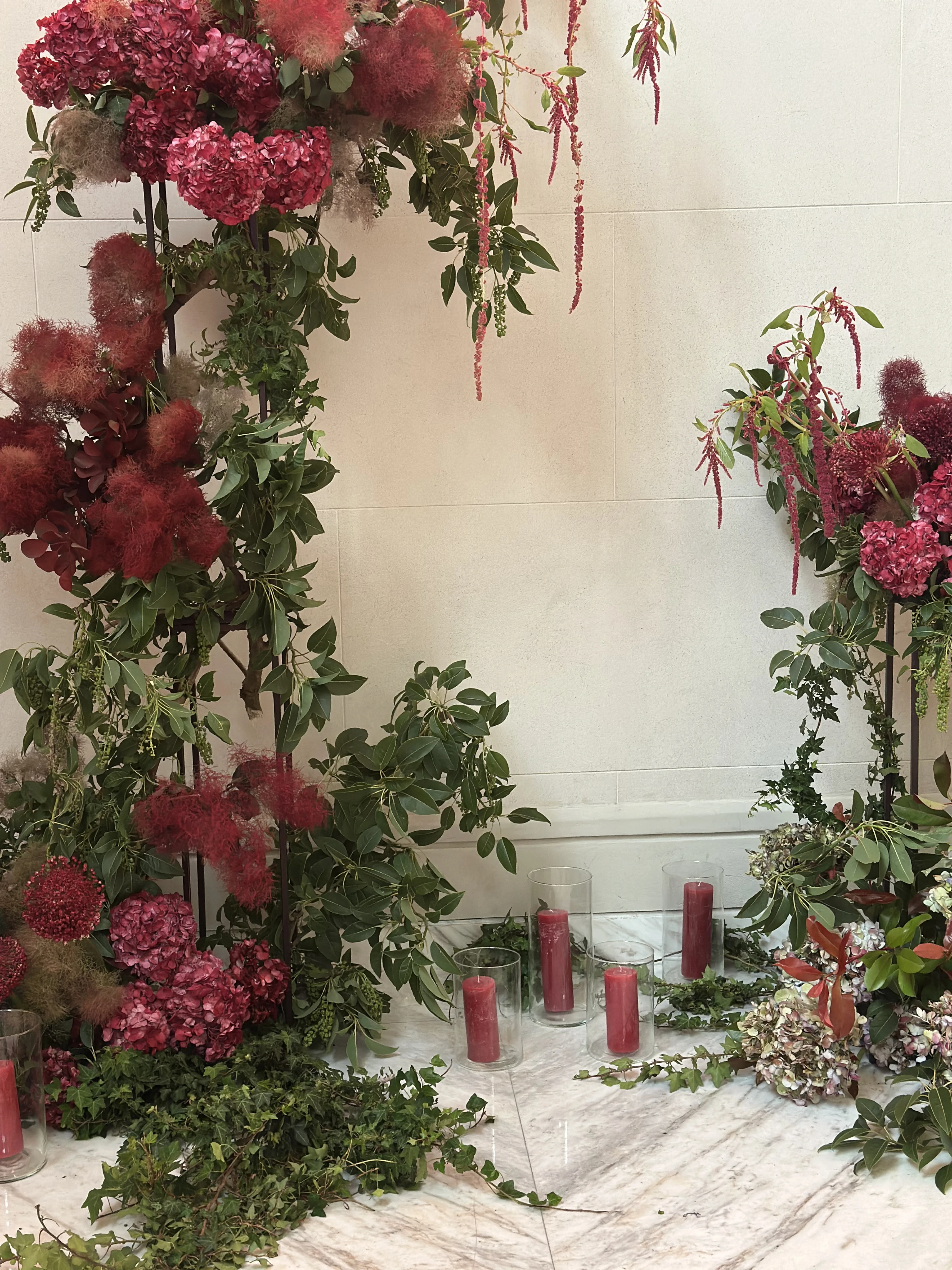 Decoración de suelo inmersiva con hortensias fucsia, follaje verde, plumeros rojos y velas en vasos de vidrio.