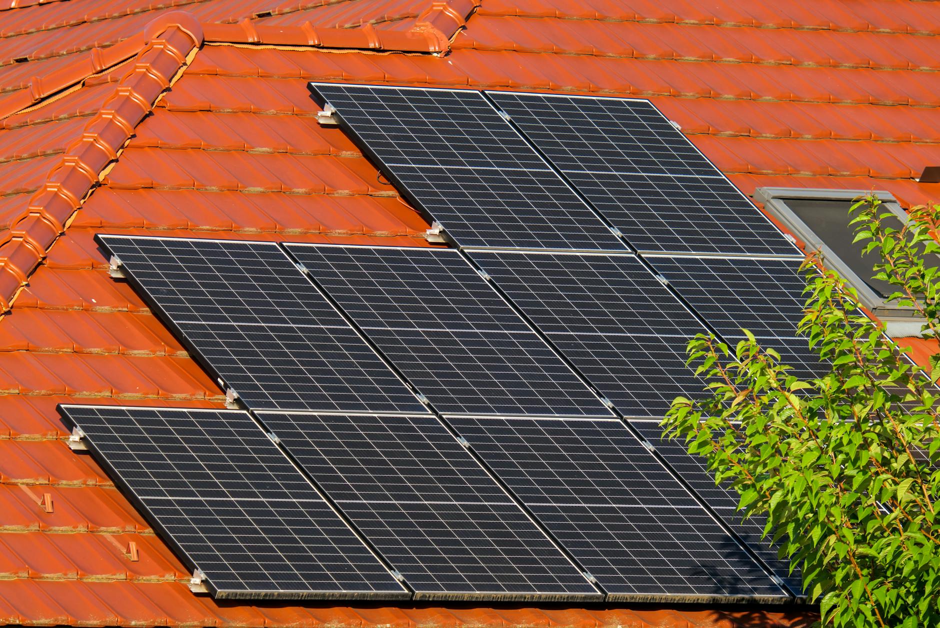 Multiple rows of solar panels mounted on a terracotta-tiled roof with a skylight and green tree foliage