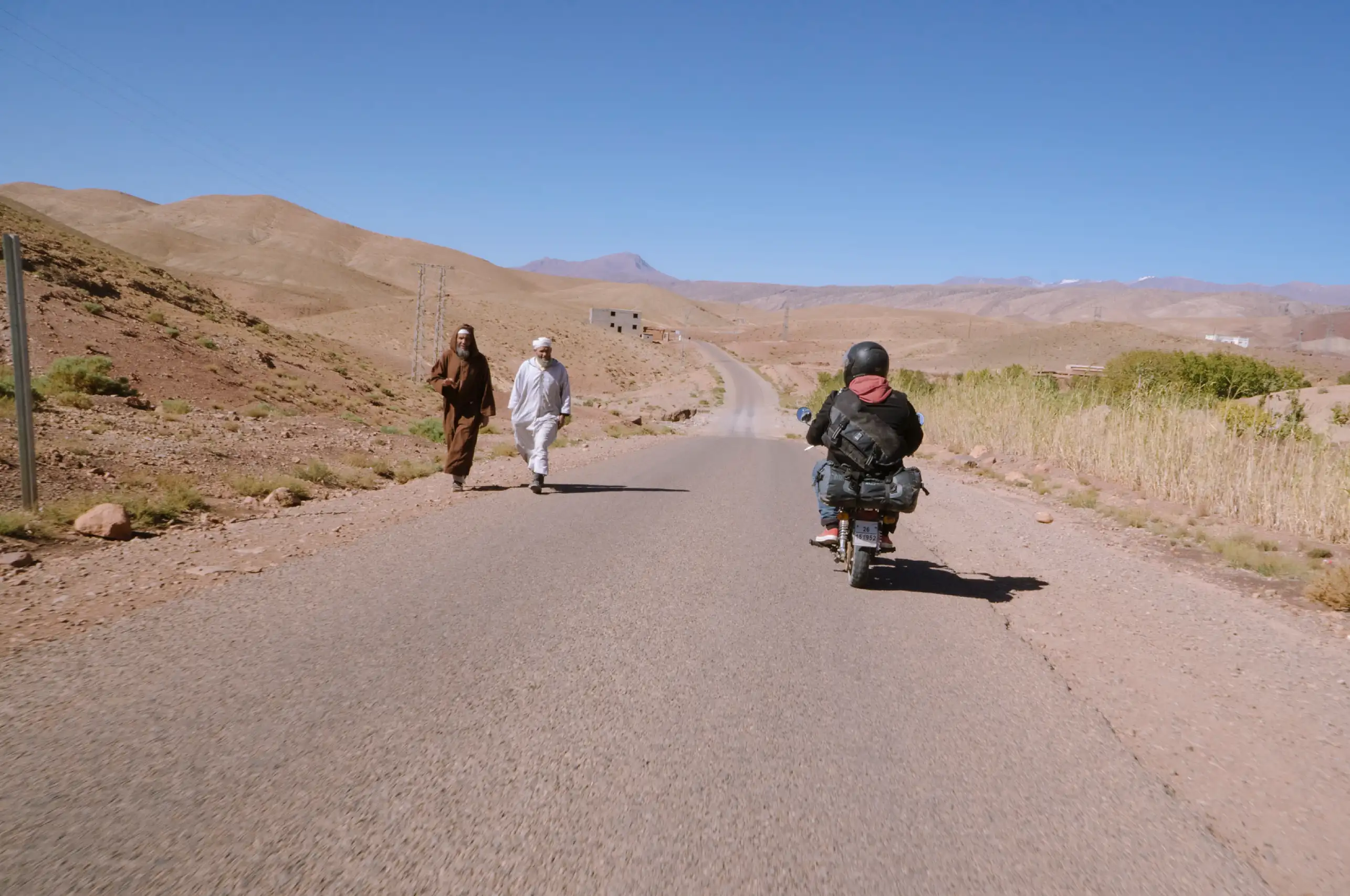 A motorcycle rider passing two men walking down a rural road in Morocco.