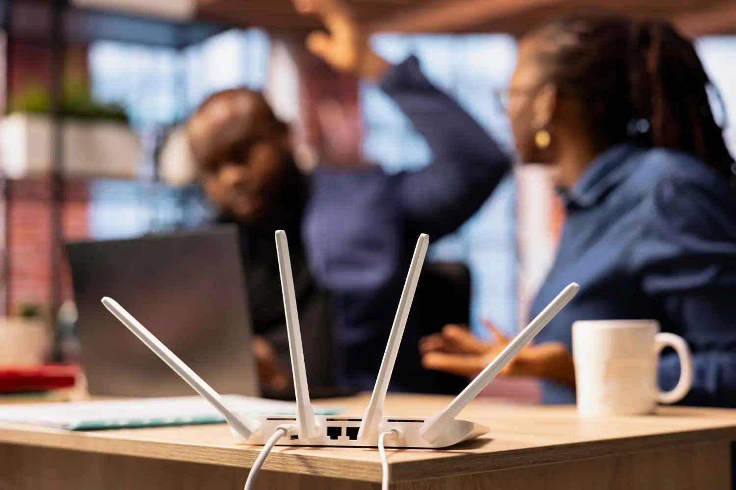 An office WiFi router in the foreground while two colleagues react to the connection going down behind it