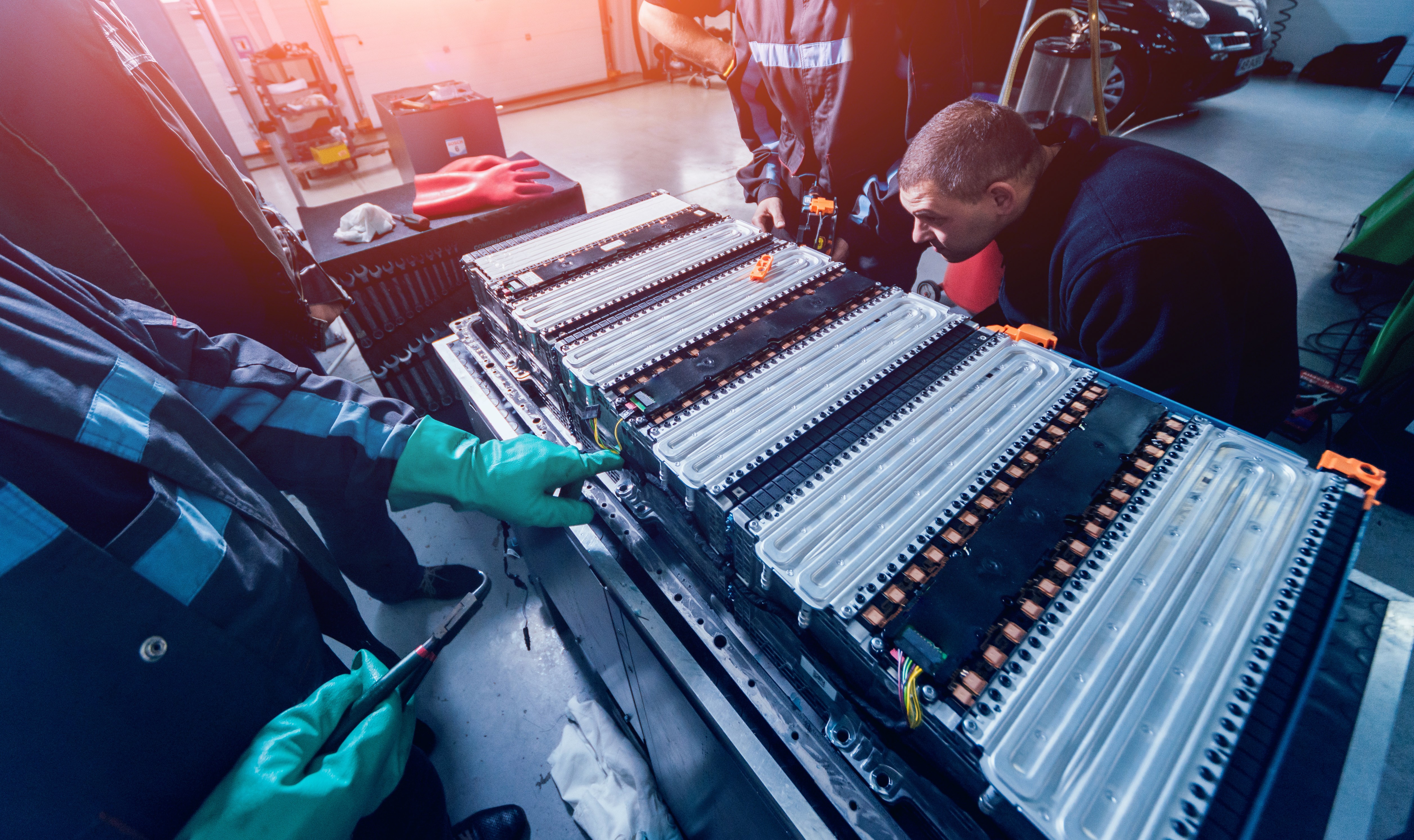 Technicians in workwear and protective gloves inspecting a large disassembled electric vehicle battery pack in a workshop