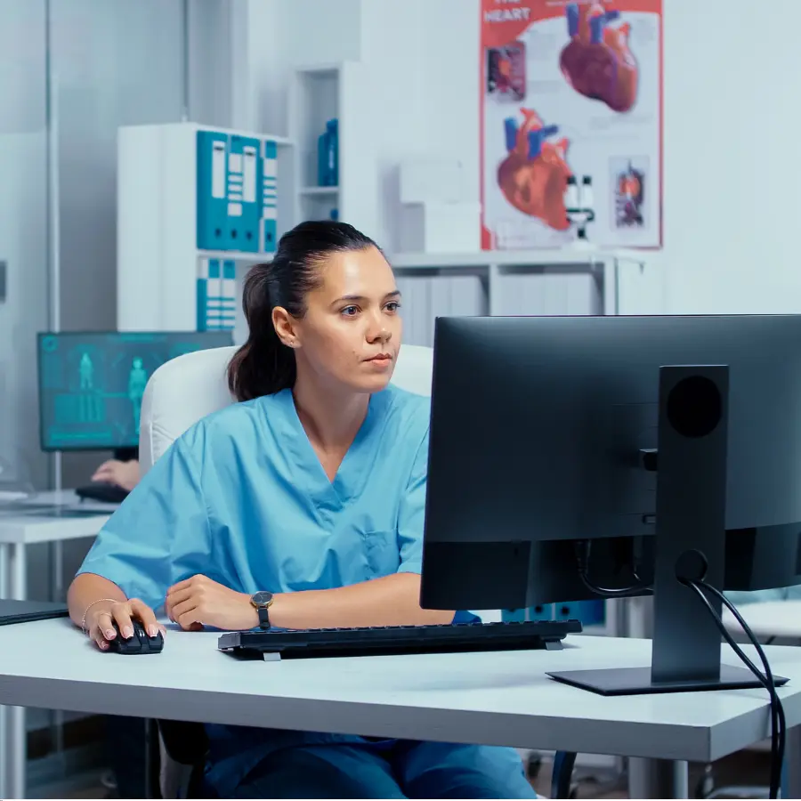 AI nurse-systems assist a focused nurse in blue scrubs as she reviews patient data on a computer in a medical office.