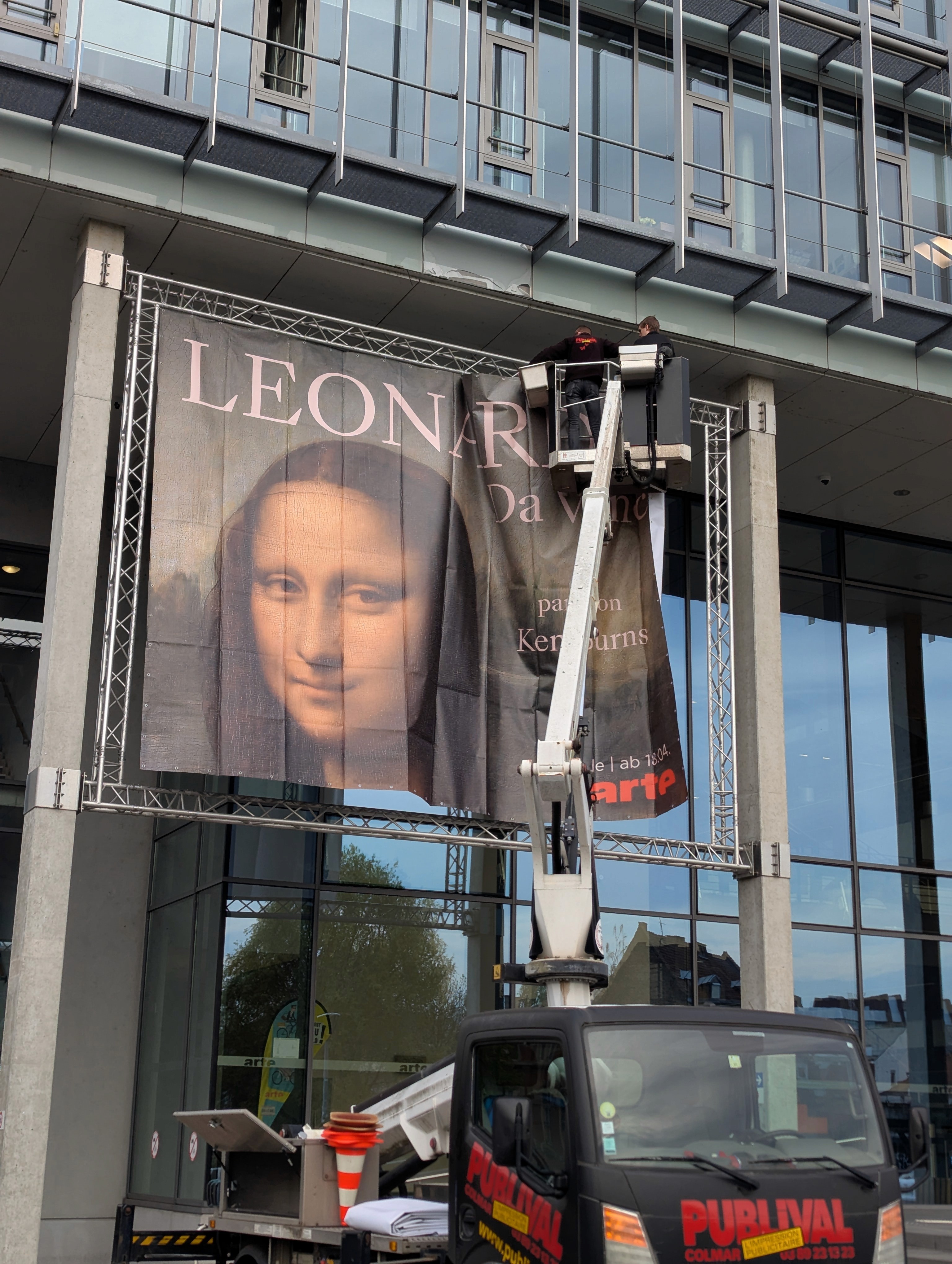 A large banner featuring a portrait of Mona Lisa is being hung on a modern building by workers in an aerial lift.