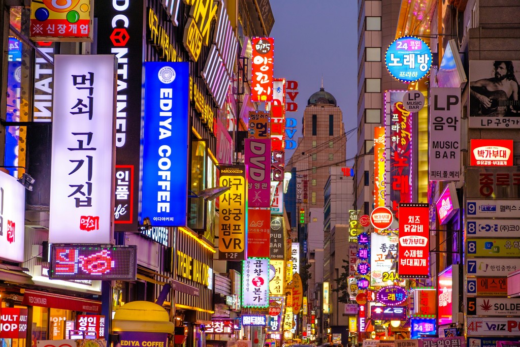 A vibrant, bustling street in Seoul, South Korea, illuminated by an array of colorful neon signs and billboards advertising coffee shops, restaurants, and entertainment venues, reflecting the energetic nightlife and commercial hub of the city.