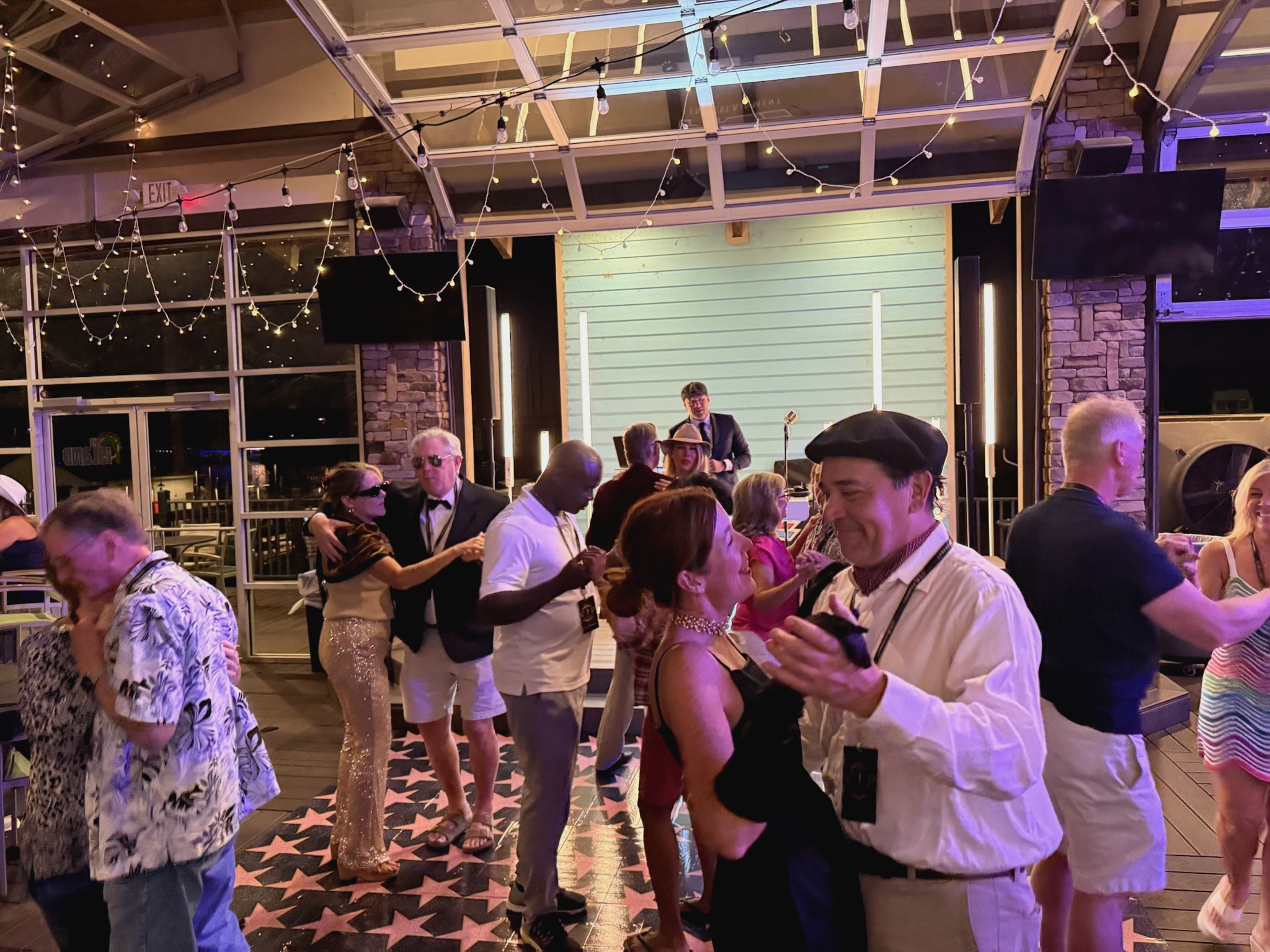 A lively dance floor features guests in elegant attire enjoying a festive indoor party, illuminated by string lights and a live music performance on stage.
