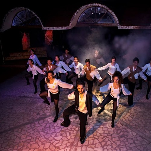 En grupp artister i matchande kostymer dansar på en scen med dimma och färgglada ljuseffekter i bakgrunden.