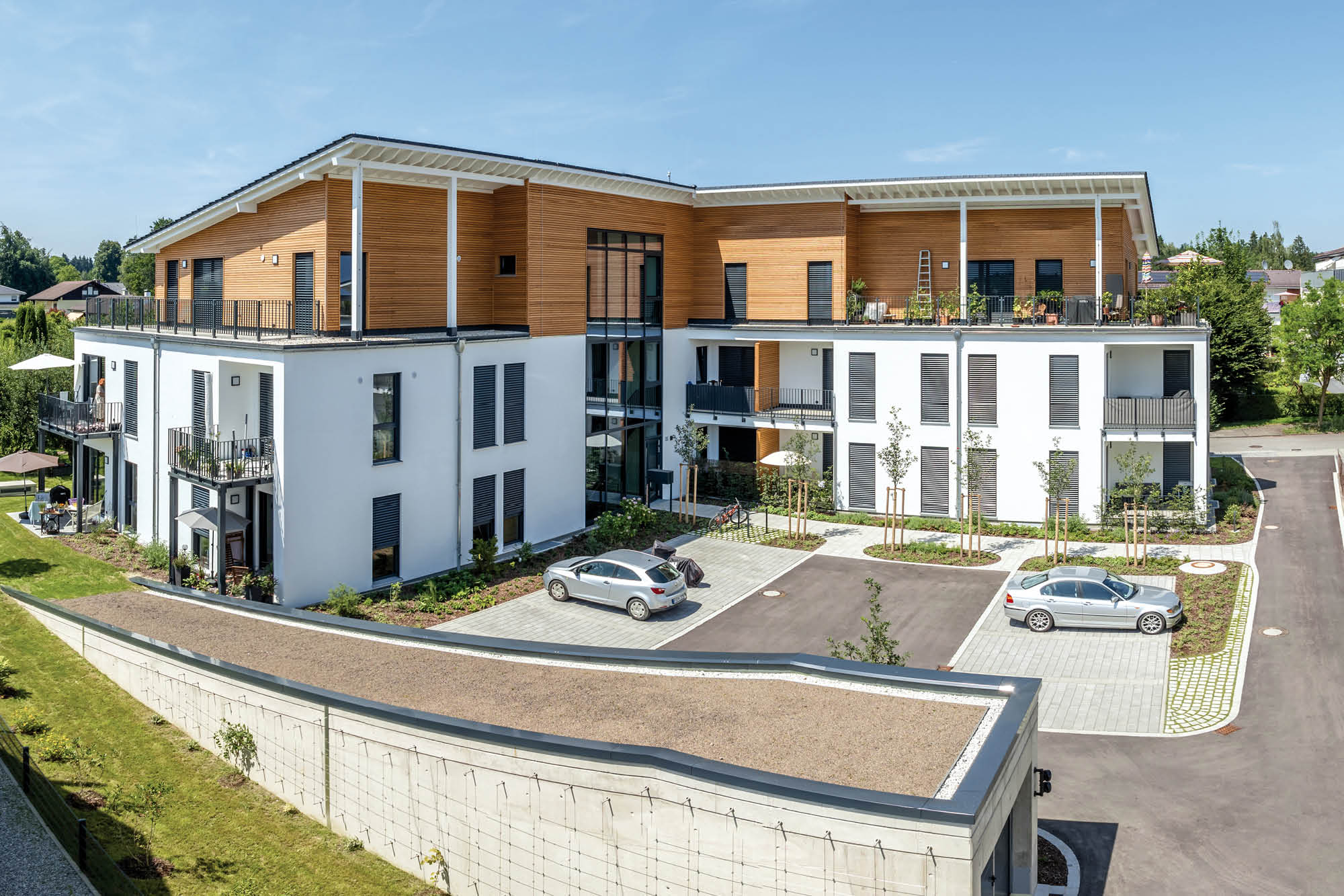 Moderne Wohnanlage mit weißer Fassade, Holzelementen und Balkonen. Zwei Autos parken auf dem gepflasterten Vorplatz.