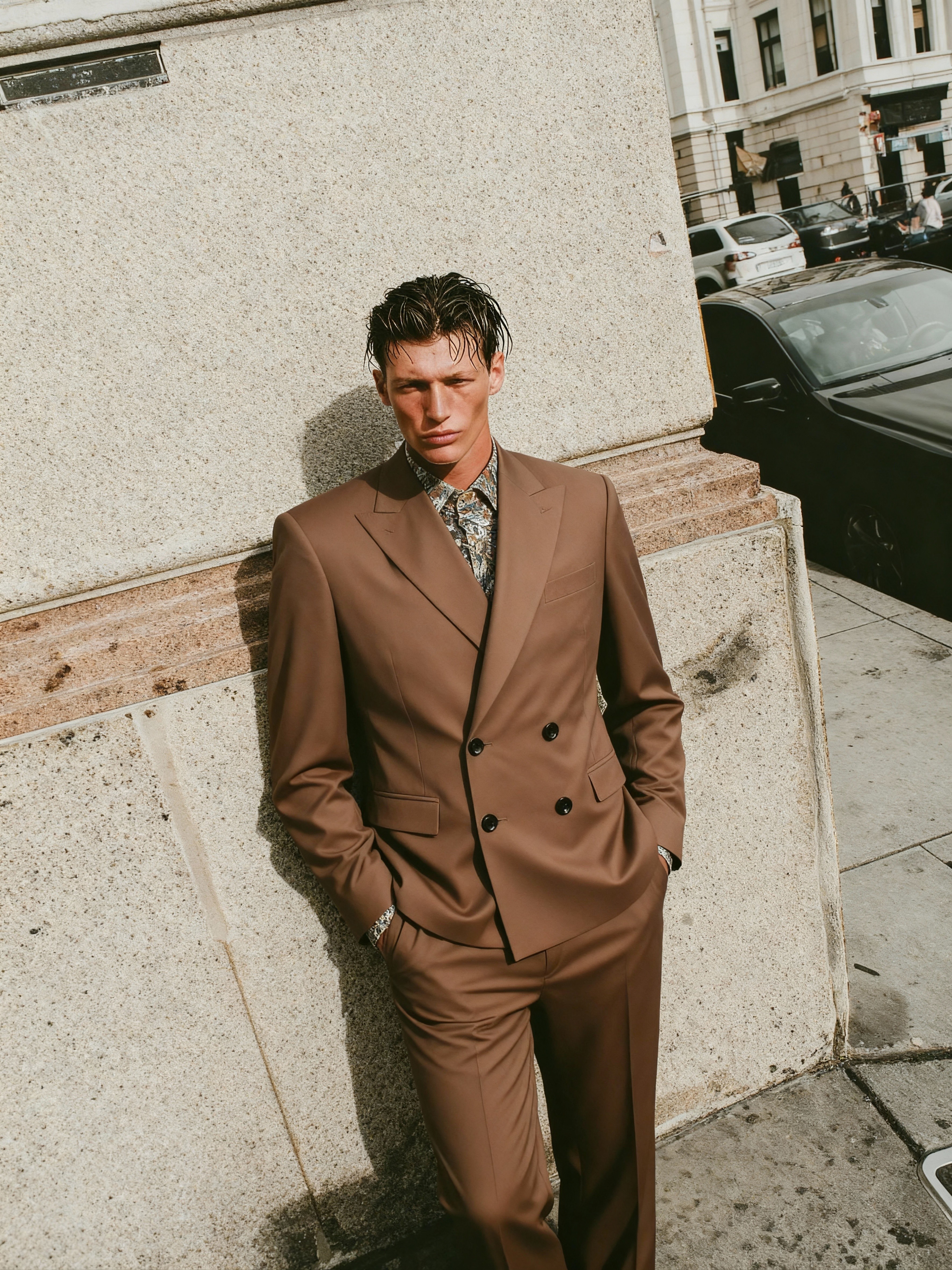 AI-generated fashion campaign image of a man in a brown double-breasted suit leaning against an urban stone wall, created by Myrovia Labs for Forzieri. AI menswear photography and luxury fashion branding.