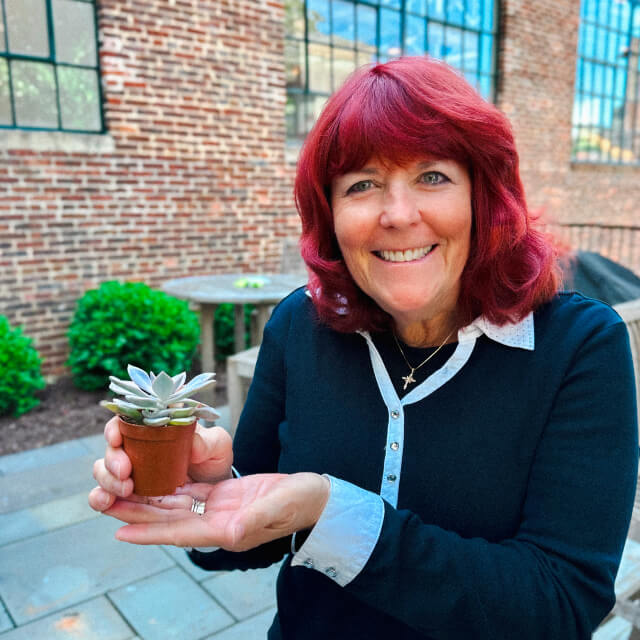 A woman with red hair smiles while holding a small potted succulent outdoors in front of a brick building, reflecting how you can grow your career in an inspiring agency environment.
