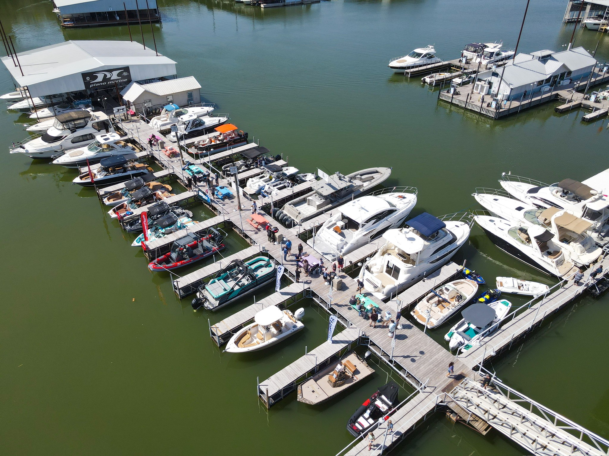 Aerial view of a bustling marina filled with various boats moored along wooden docks, surrounded by green-tinged water, with people walking around and nearby dockside buildings.