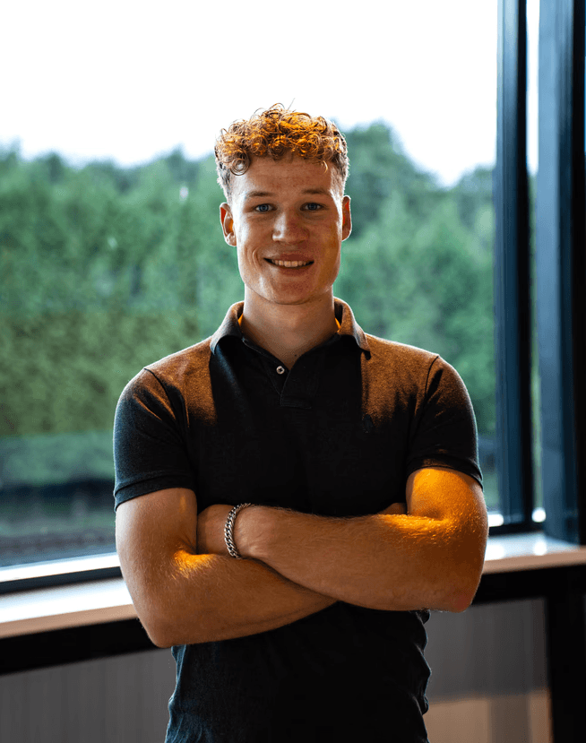 A smiling young man with crossed arms, wearing a plaid shirt and white t-shirt, poses against a dark background.