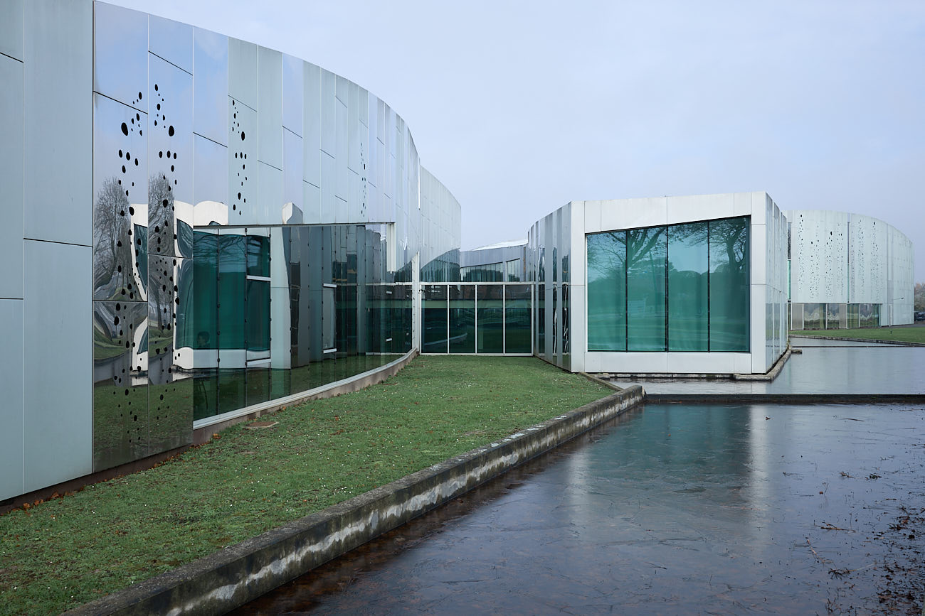 Vue Est d'architecture extérieure du Centre Culturel de Lesquin : façade vitrée et miroir avec des trous rond formant des bulles, se reflétant entre un bassin d'eau gelé et une partie d'herbe entretenue.