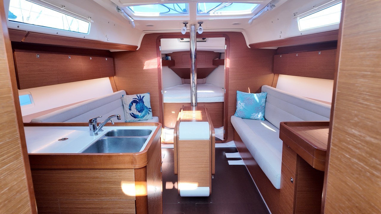 Interior view of the Dufour 350 GL's saloon looking forward toward the V-berth, with gray cushioned settees on either side of a fold-out teak table, overhead hatches, and a mast compression post at center.