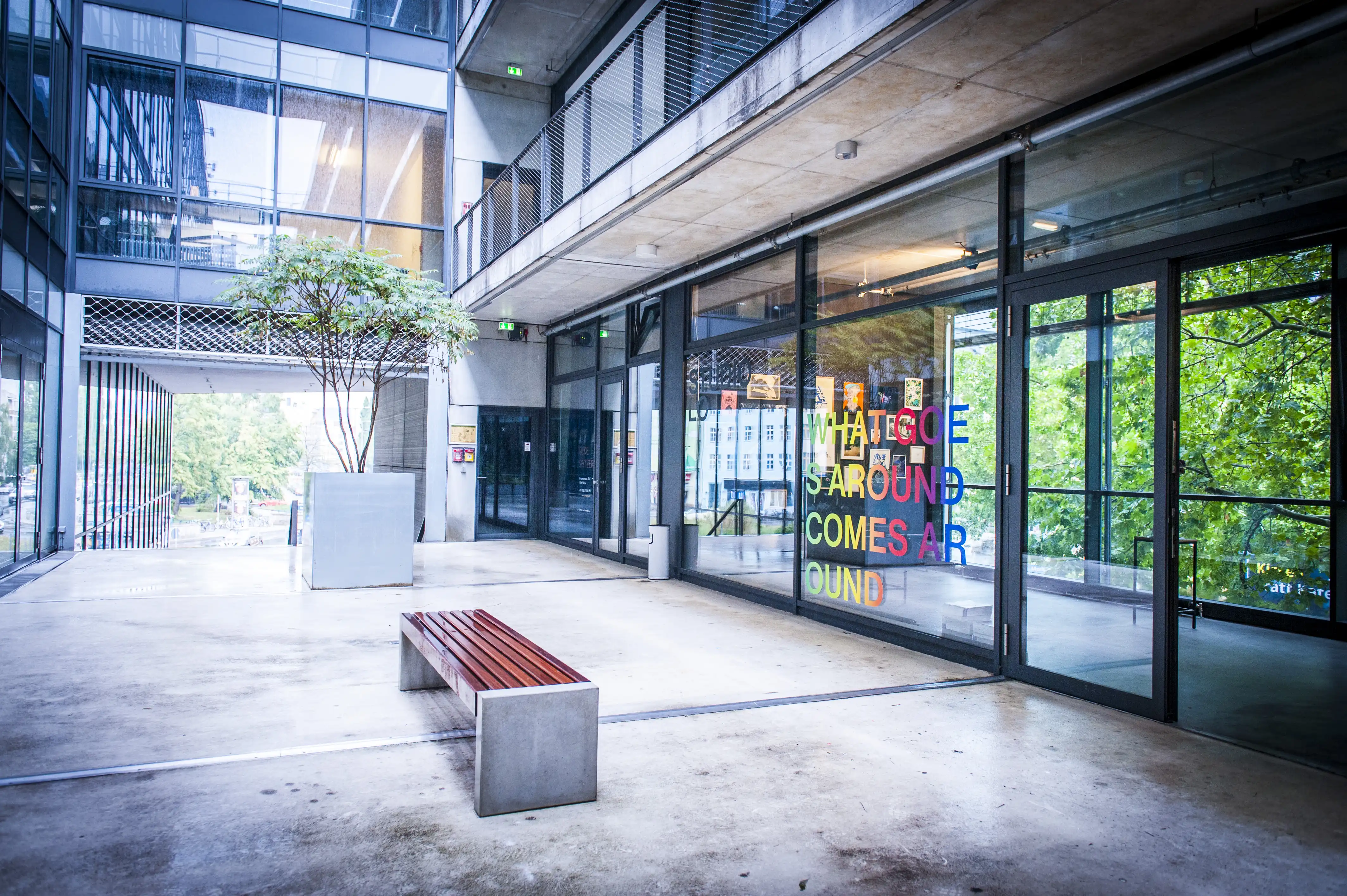Courtyard view of the STUDIOLO gallery with colorful What Goes Around Comes Around lettering on the glass facade and a wooden bench in front.