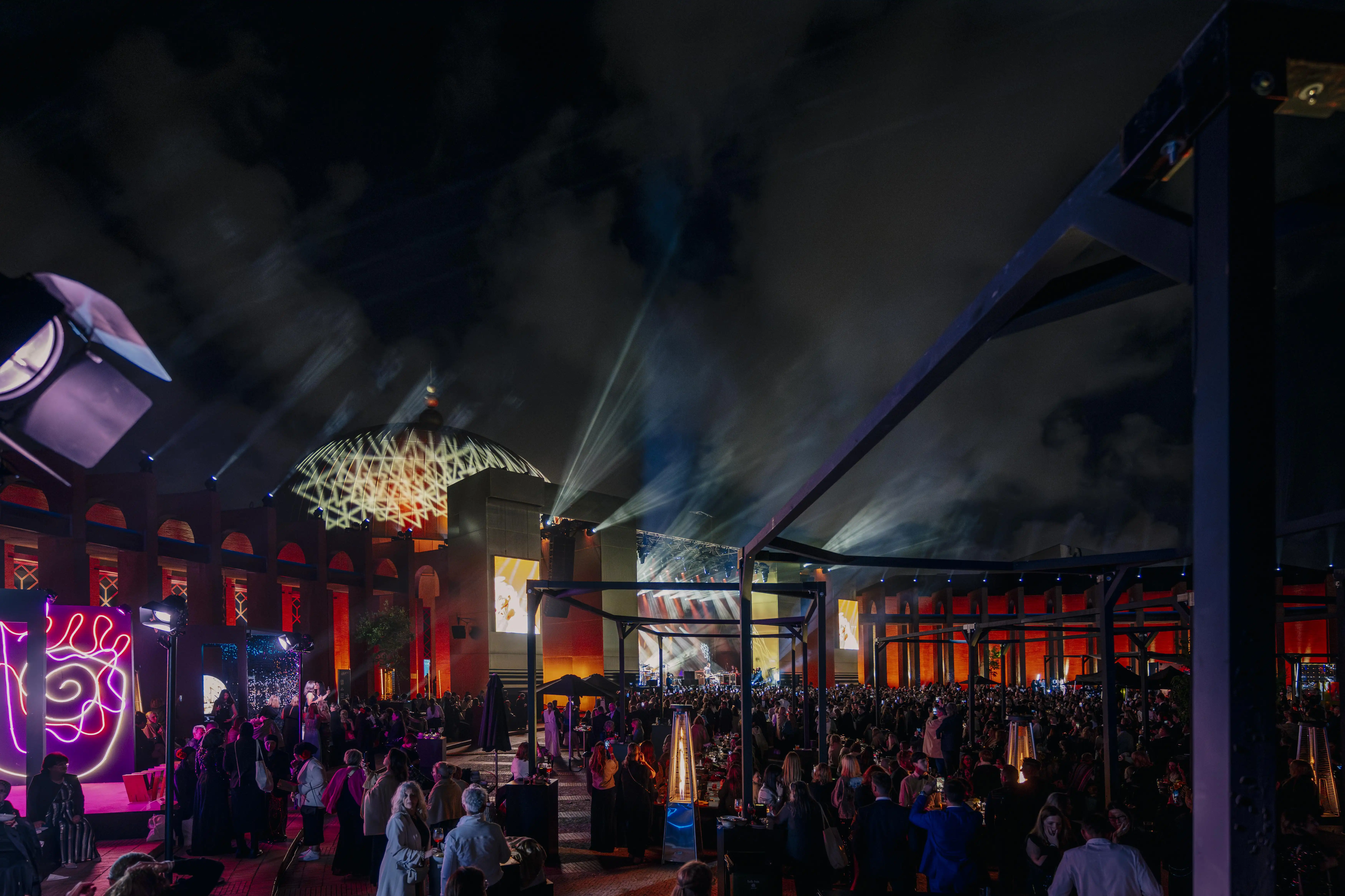 Night-time outdoor area with crowd, light beams and red-lit historic facades in Seville.