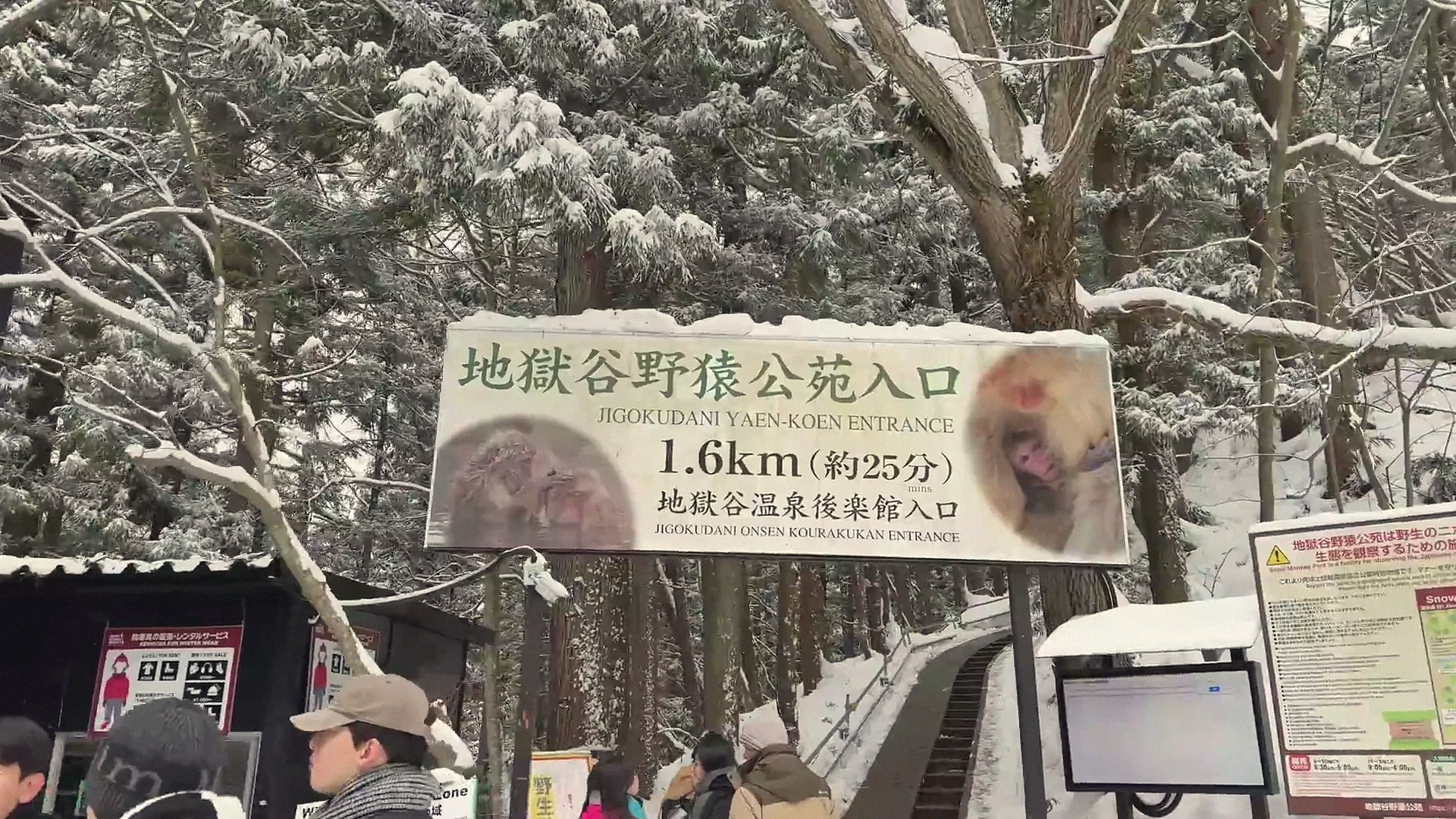 The bilingual entrance banner for Jigokudani Yaen-kōen — '地獄谷野猿公苑入口 / JIGOKUDANI YAEN-KOEN ENTRANCE, 1.6km (約25分)' — as visitors pass beneath in snow.