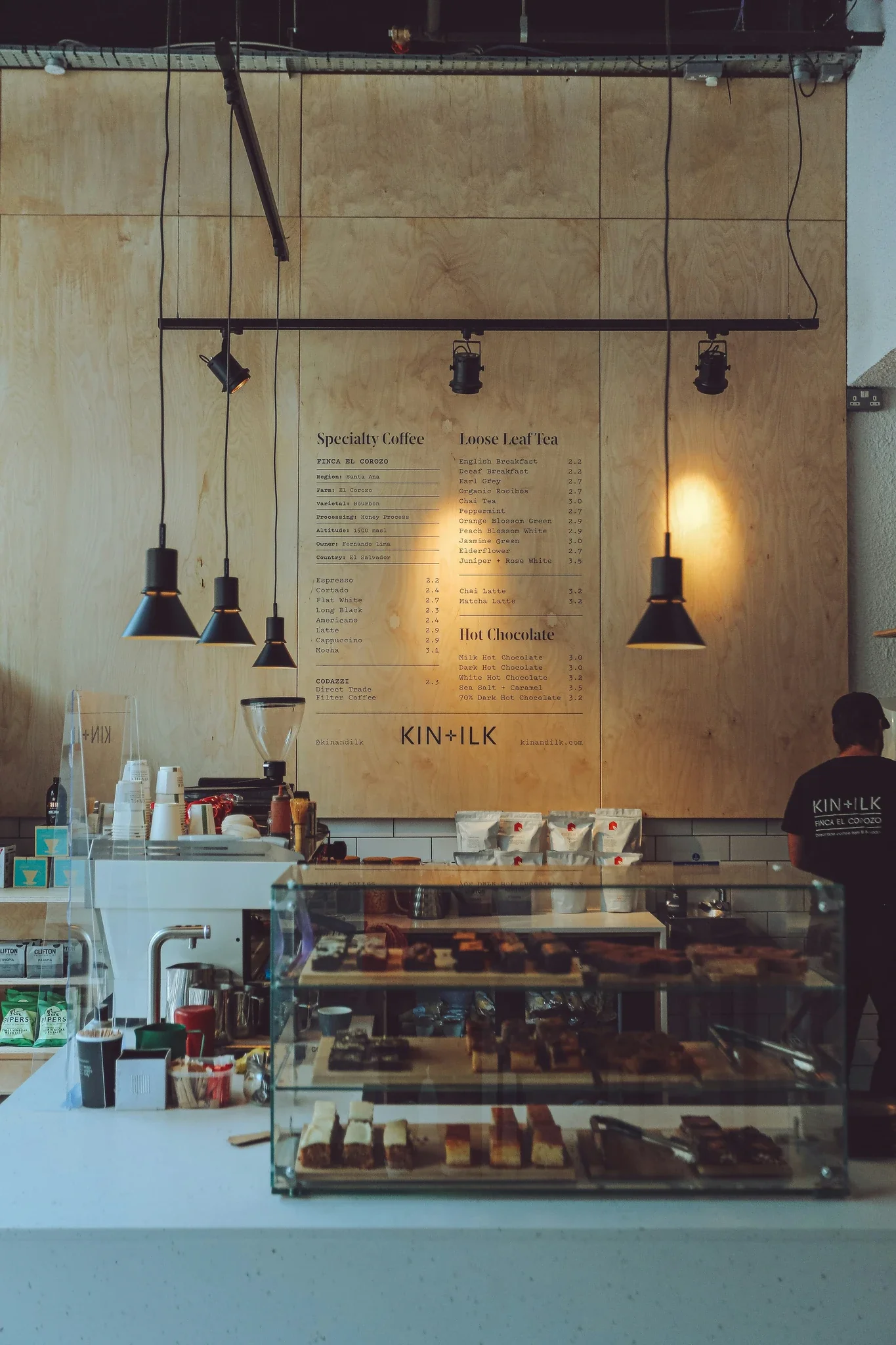 Modern café interior with "KIN+ILK" menu board displaying specialty coffee, loose leaf tea, and hot chocolate options above counter with pastry display.