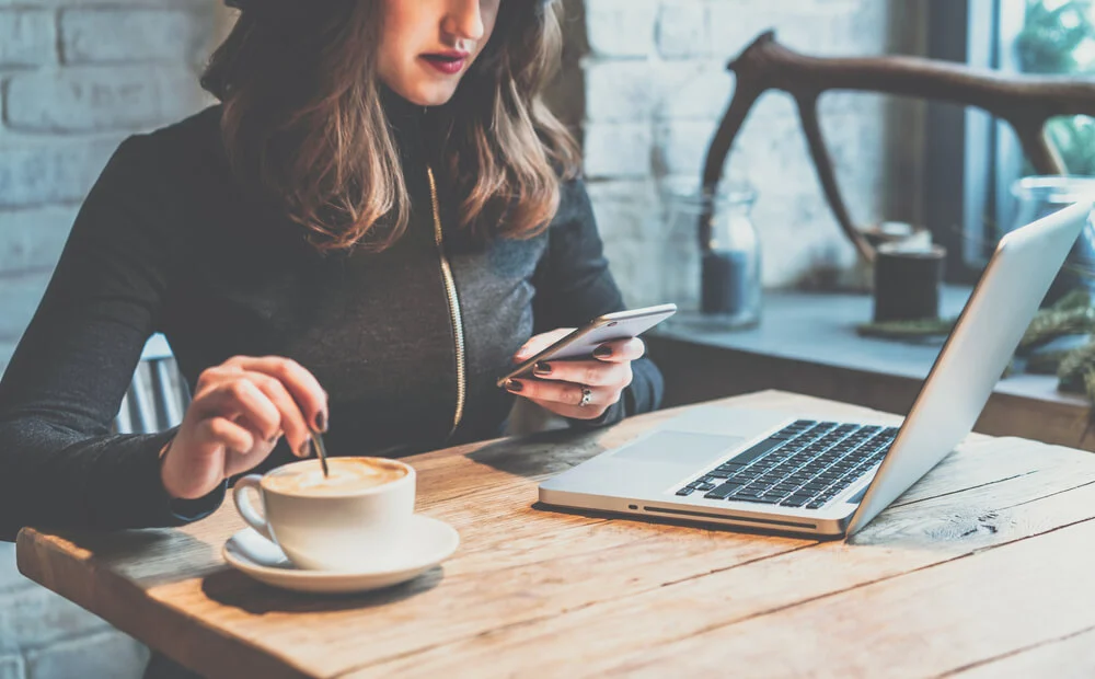 Woman in a café using a smartphone while stirring coffee beside an open laptop
