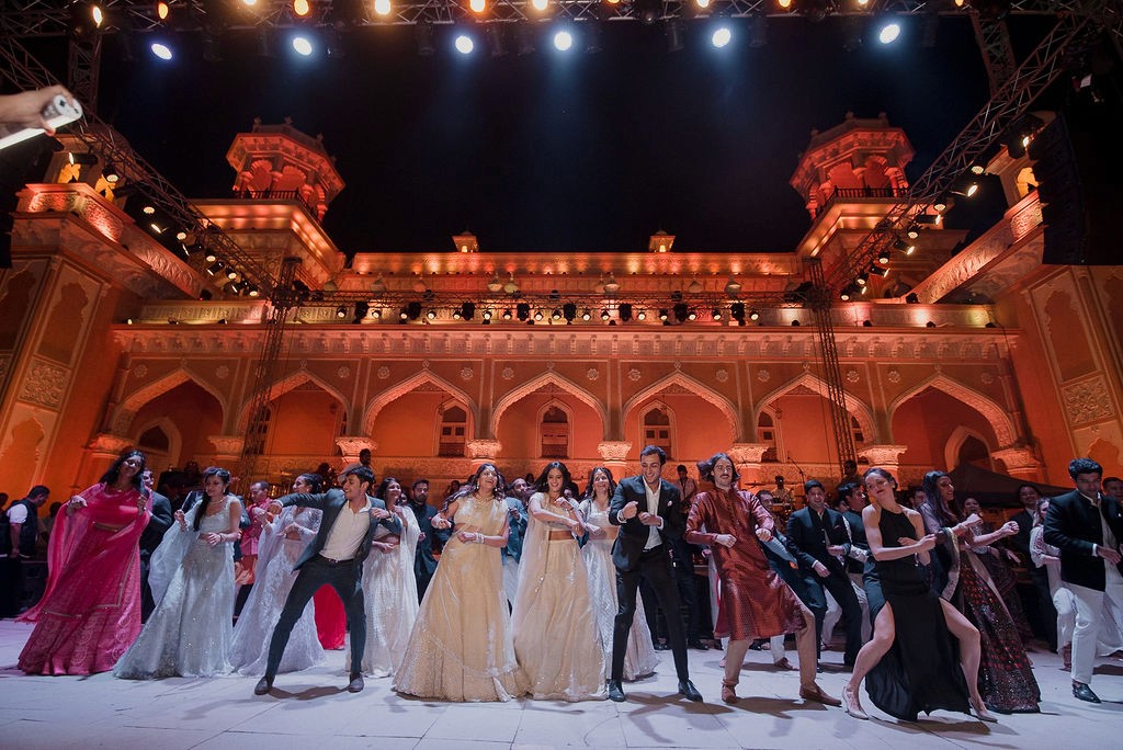 A rustic outdoor market stall displays an array of fresh produce including vibrant tomatoes, leafy greens, and a variety of olives, set against the backdrop of a picturesque rural Italian village.