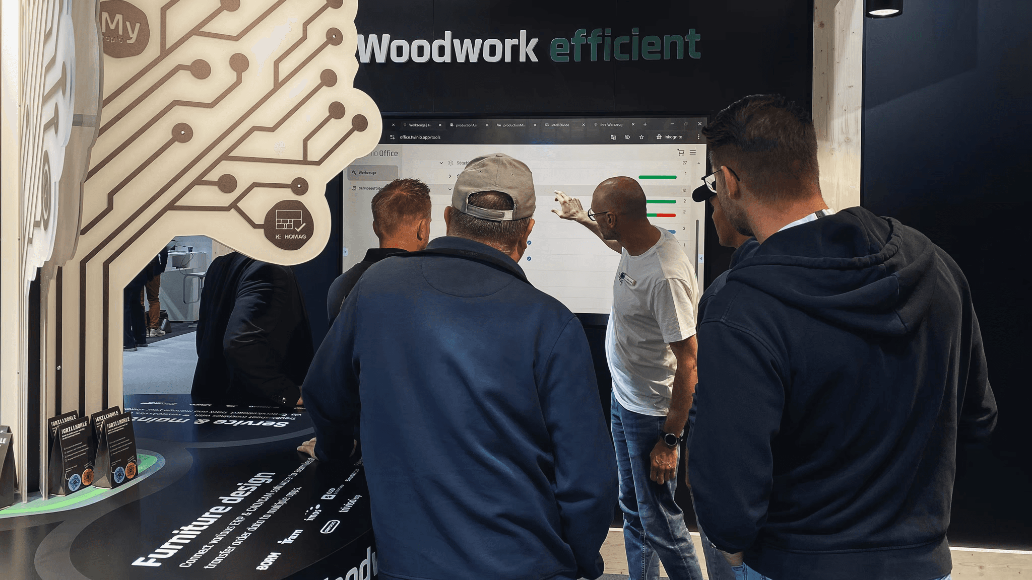 Person zeigt auf einem messestand auf einen Bildschirm während Leute zuschauen